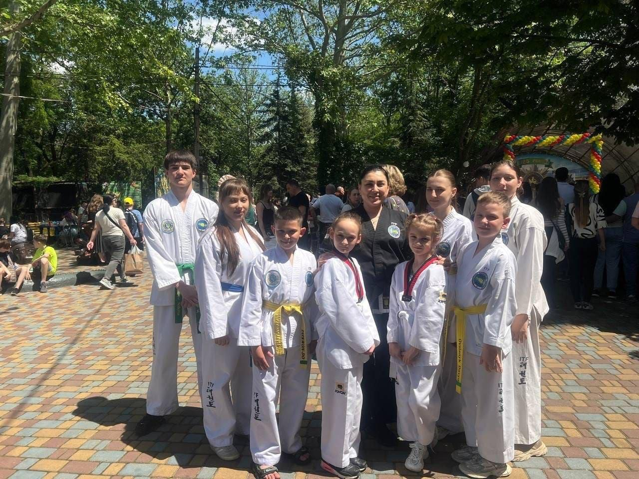 Група з восьми юних спортсменів різного віку у білих кімоно (добоках) для тхеквондо та їхня усміхнена жінка-тренер у чорній формі позують для спільного фото на брукованій алеї парку в межах Тижня спорту, організованого миколаївським координаційним центром «Вільні разом» для переселенців із Херсонської області. Діти із жовтими, зеленими та синіми поясами вишикувалися в один ряд на передньому плані, а наставниця тепло обіймає їх за плечі на тлі соковитої літньої зелені дерев, святкової арки з різнокольоровими повітряними кульками та відпочивальників у парку.
