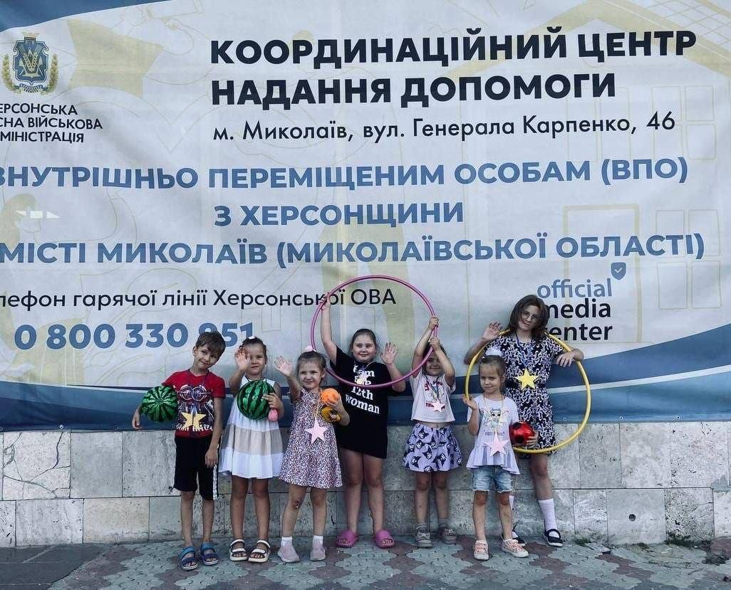 Шість дітей та одна жінка, яка частково потрапила в кадр ліворуч, позують на тлі великого офіційного банера координаційного центру «Вільні разом» у місті Миколаїв під час спортивних змагань «Веселі старти», що відбулися в межах Тижня спорту для переселенців із Херсонщини. Разом на знімку зафіксовано сім людей — шість радісних дітей у літньому одязі в центрі та доросла жінка, від якої видно руку й плече з лівого краю кадру. Хлопчик і дівчинка тримають у руках два великі смугасті кавуни, інші дівчатка весело підняли руки, тримаючи різнокольорові обручі-хулахупи та золоту зірку на паличці, святкуючи спортивні вихідні на тлі банера з адресою хабу.