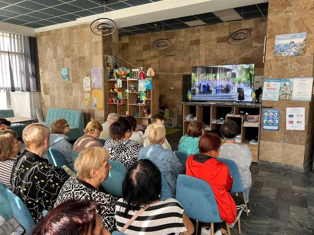 Група людей похилого віку та дорослих переселенців із Херсонщини сидить на блакитних стільцях у залі миколаївського координаційного центру «Вільні разом» і дивиться онлайн-трансляцію святкового концерту «У серці Україна, у пісні — душа!» до Дня української музики. Глядачі повернуті спиною до камери та уважно спостерігають за виступом хору на великому телевізійному екрані, встановленому на тумбі по центру кімнати. Захід проходить у світлому приміщенні з бежевими плитковими стінами, де ліворуч облаштовано стелажі з дитячими виробами та блакитний диван, а праворуч на колоні розвішані інформаційні плакати й оголошення для відвідувачів хабу.