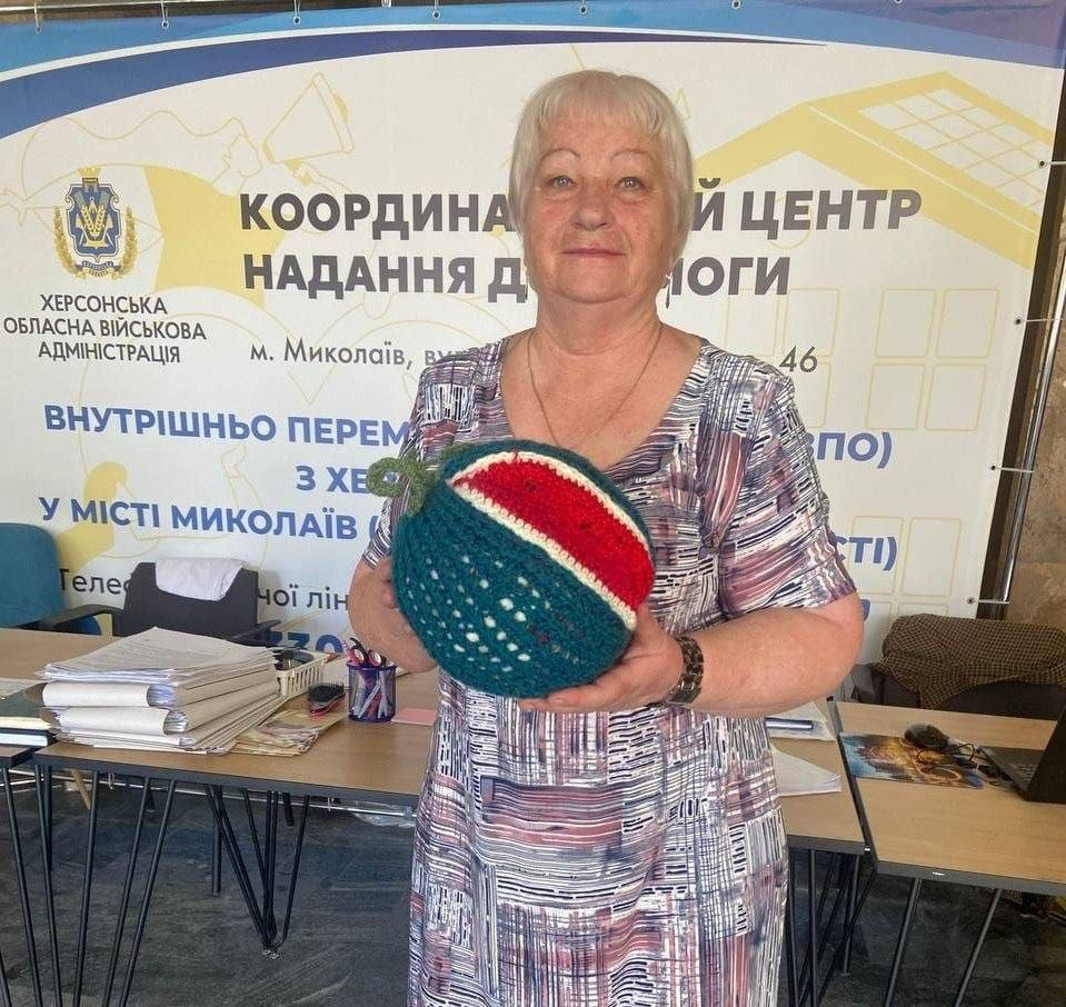 Жінка похилого віку зі світлим волоссям у кольоровій сукні стоїть у приміщенні хабу і тримає в руках оригінальний в'язаний виріб у формі скибки кавуна, виготовлений під час творчого майстер-класу від ГО «Дівчата» з нагоди 248-річчя Херсона, організованого миколаївським координаційним центром «Вільні разом» для переселенців із Херсонщини. Учасниця заходу посміхається в камеру на тлі великого офіційного банера з написом «КООРДИНАЦІЙНИЙ ЦЕНТР НАДАННЯ ДОПОМОГИ ВНУТРІШНЬО ПЕРЕМІЩЕНИМ ОСОБАМ (ВПО) З ХЕРСОНЩИНИ В МІСТІ МИКОЛАЇВ» та гербом Херсонської ОВА. На задньому плані помітно робочі столи з ноутбуком, паперами, канцелярією та пачками документів.