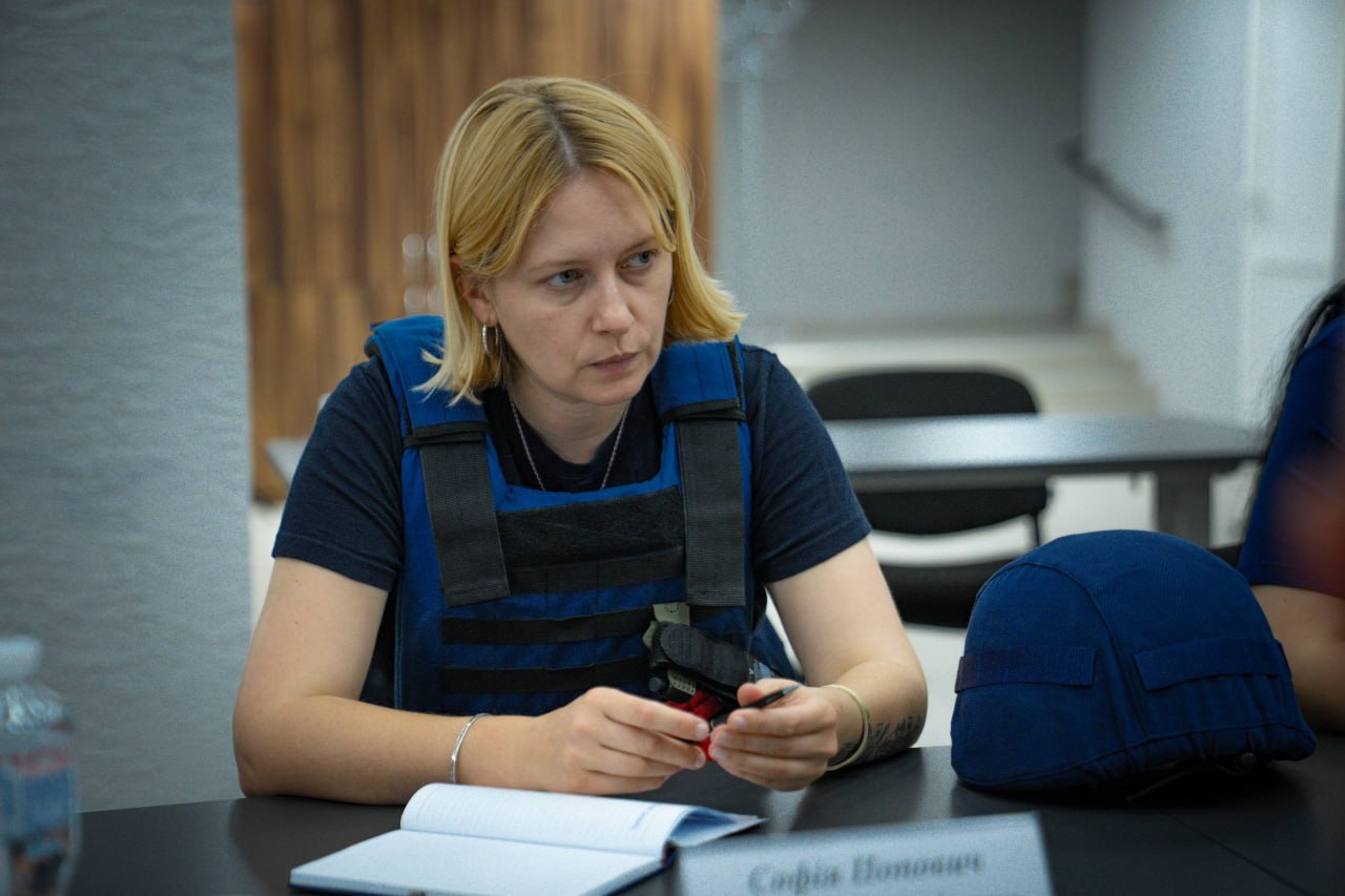 Виконуюча обов’язки директора чеської гуманітарної організації «Людина в біді» (People in Need) в Україні Софія Попович сидить за столом під час зустрічі з представниками Херсонської ОВА щодо підготовки деокупованих громад до опалювального сезону. Керівниця у синьому захисному бронежилеті поверх чорної футболки тримає в руках ручку, а перед нею на столі лежить відкритий блокнот для записів та табличка з її ім'ям. Праворуч на темній поверхні столу розташована синя захисна каска, а на розмитому задньому плані помітно інтер'єр офісного приміщення, де учасники погоджують залучення ресурсів міжнародних партнерів для підтримки прифронтових територій.