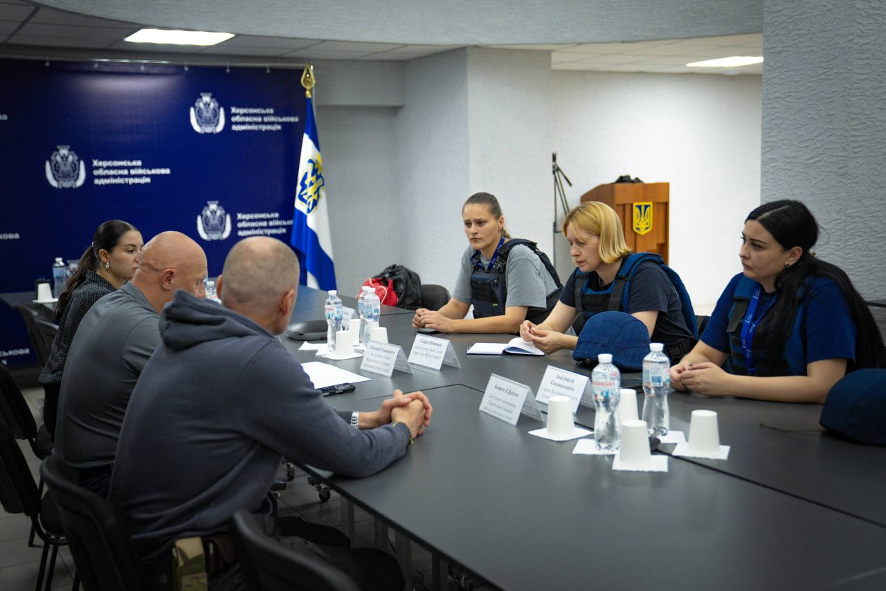 Представники Херсонської ОВА та чеської гуманітарної організації «Людина в біді» (People in Need) за круглим столом обговорюють підготовку деокупованих громад до зими. На передньому плані ліворуч, боком та спиною до камери, сидить керівництво області: заступник начальника Херсонської МВА Антон Єфанов у темно-синьому худі, заступник начальника Херсонської ОВА Євгеній Ігнатенко в сірому поло (у центрі групи) та начальниця управління міжнародного співробітництва Марія Богодухова. Навпроти них, у синій фірмовій формі, розмістилися чеські партнери: найближче до глядача ліворуч сидить представниця організації в бронежилеті, по центру — виконуюча обов’язки директора «People in Need» в Україні Софія Попович, також у захисному спорядженні, а праворуч — їхня колега. Зустріч, присвячена координації опалювальної допомоги та забезпеченню жителів прифронтових територій твердим паливом, проходить у залі засідань на тлі офіційного темно-синього банера та прапора Херсонської області на флагштоці в кутку кімнати.