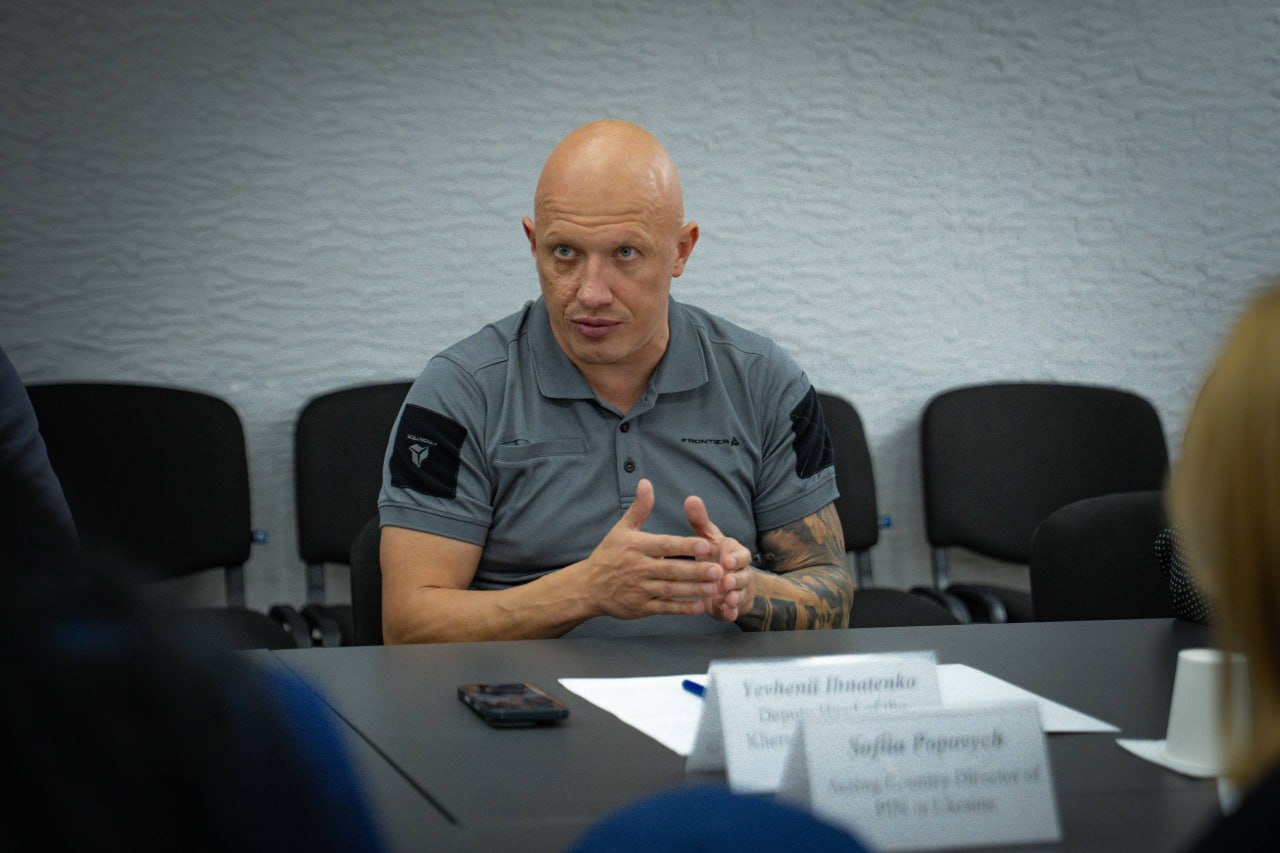 Заступник начальника Херсонської ОВА Євгеній Ігнатенко сидить за робочим столом по центру кадру під час зустрічі з представниками чеської організації «Людина в біді» (People in Need) щодо підготовки громад до опалювального сезону. Чоловік у сірому спортивному поло з тактичними нашивками на рукавах активно жестикулює руками, пояснюючи потреби прифронтових територій у твердому паливі. Перед ним на столі лежать папери, ручка, мобільний телефон та інформаційні таблички. На задньому плані видно світлу фактурну стіну та ряд порожніх чорних офісних стільців.