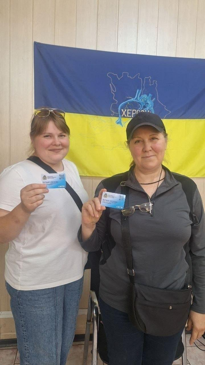 Дві жінки з числа переселенців із Херсонщини стоять у приміщенні одеського координаційного центру «Вільні разом» і тримають у руках отримані пластикові картки в межах реалізації проєкту «Картка Херсонщини». Ліворуч стоїть молода усміхнена жінка у білій футболці, блакитних джинсах та з сонцезахисними окулярами на голові, а праворуч від неї — жінка у сірій кофті, темній кепці та з чорною сумкою через плече. Обидві учасниці повернуті обличчям до камери і демонструють свої синьо-блакитні картки на тлі світлих стін кімнати та великого синьо-жовтого прапора з контурами Херсонської області й написом «ХЕРСОН», де триває системна видача посвідчень та розширення партнерської мережі.