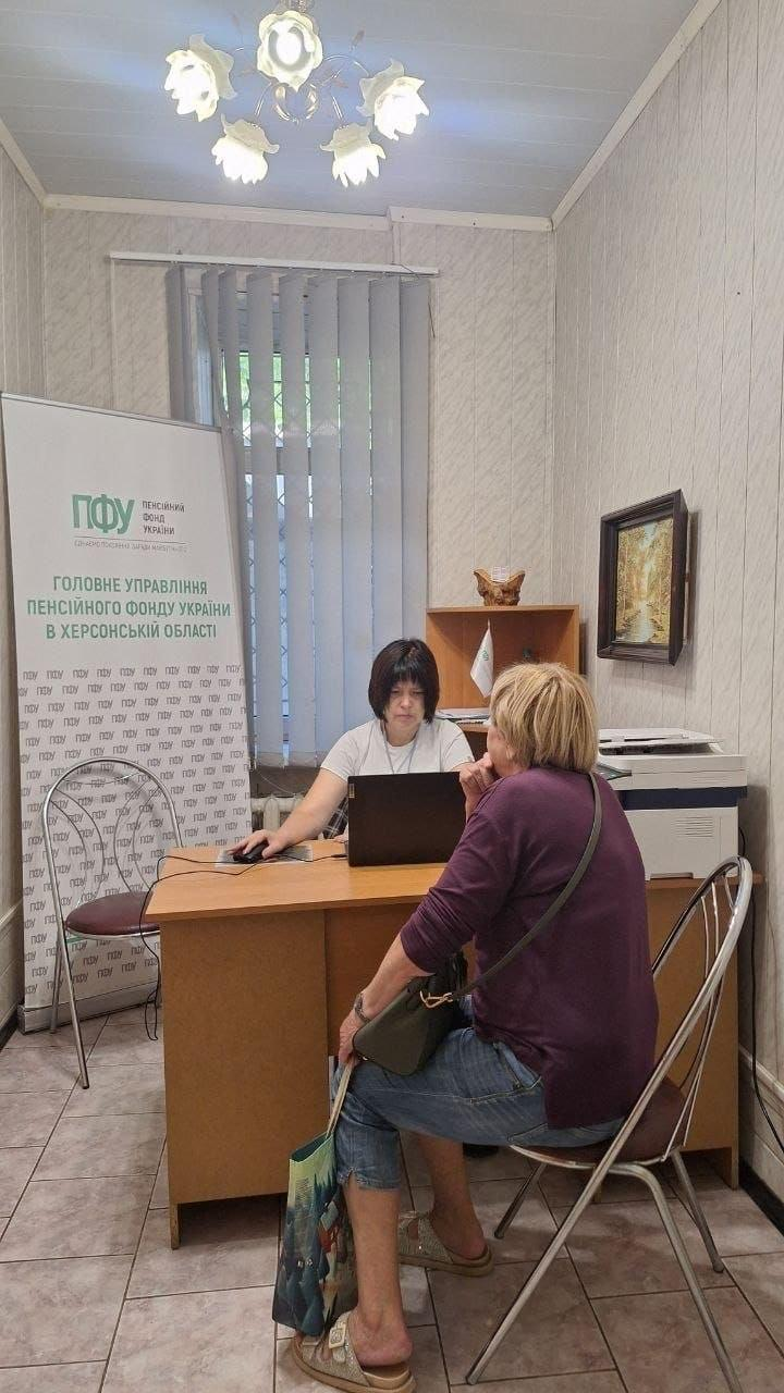 Фахівець Головного управління Пенсійного фонду України в Херсонській області консультує жінку з числа переселенців у кабінеті одеського координаційного центру «Вільні разом» під час прийому громадян. Співробітниця з короткою темною стрижкою в білій футболці сидить за робочим столом із відкритим ноутбуком Lenovo, а перед нею боком до камери на металевому стільці розмістилася відвідувачка у фіолетовій кофті та блакитних джинсових капрі з сумкою через плече, яка прийшла для вирішення пенсійних питань. Ліворуч біля столу встановлено великий вертикальний ролап-банер із логотипом «ПФУ» та написом «ГОЛОВНЕ УПРАВЛІННЯ ПЕНСІЙНОГО ФОНДУ УКРАЇНИ В ХЕРСОНСЬКІЙ ОБЛАСТІ», а праворуч на тумбі розташований великий офісний принтер під світлою люстрою на п'ять плафонів, де протягом дня фахівець ПФУ успішно прийняв 8 громадян.