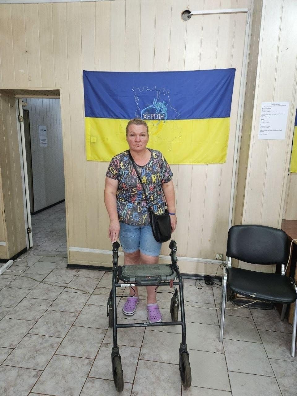 Жінка середнього віку у різнокольоровій футболці та блакитних джинсових шортах спирається на нові чотириколісні ходунки-ролатор, які вона отримала як допоміжний засіб реабілітації в одеському координаційному центру «Вільні разом» від організації «Фонд громади міста Херсон «Захист»». Переселенка з Херсонщини стоїть у приміщенні хабу, тримаючи руки на ручках темних ходунків, на тлі закріпленого на стіні великого синьо-жовтого прапора з написом «ХЕРСОН». Учасниця заходу повернута обличчям до камери, а праворуч від неї в кімнаті з кахельною підлогою помітно чорний офісний стілець та інформаційний листок, де хаб системно допомагає громадянам з обмеженими фізичними можливостями.