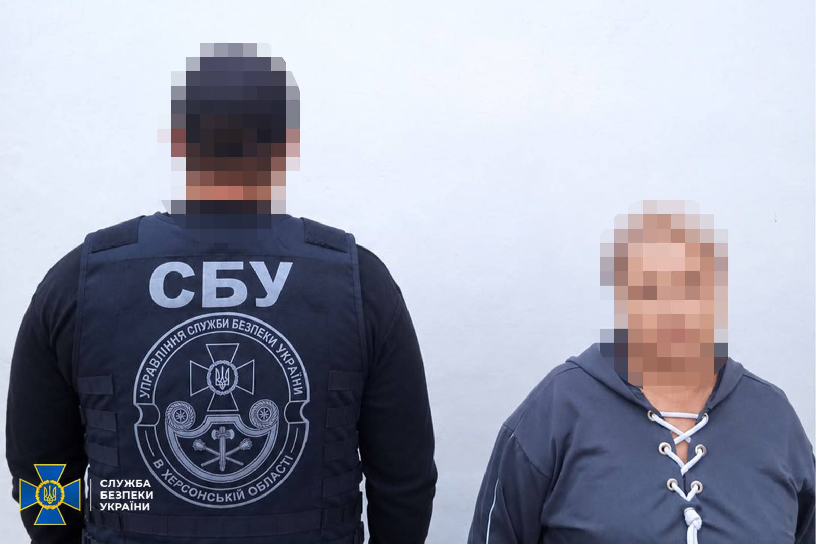 Співробітник Управління СБУ в Херсонській області стоїть спиною до камери поруч із затриманою мешканкою Бериславського району, яка брала участь в організації незаконного референдуму та намагалася втекти до Німеччини. Ліворуч у кадрі зображений офіцер у чорній кофті та тактичному жилеті з великим білим написом «СБУ» та круглим шевроном «Управління Служби безпеки України в Херсонській області» на тлі світлої стіни. Праворуч від нього обличчям до камери стоїть затримана колаборантка у темно-сірій кофті на білій шнурівці біля коміра. Обличчя жінки та потилиця співробітника спецслужби приховані піксельним розмиттям, а в лівому нижньому кутку знімка розміщено офіційний логотип Служби безпеки України.