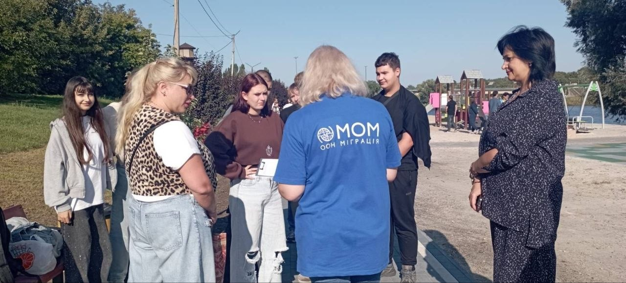 Група переселенців із Верхньорогачицької громади та організаторів спілкується на відкритому повітрі під час екологічного заходу на території міського пляжу «Райдуга» в Первомайську, організованого за підтримки Міжнародної організації з міграції (МОМ). По центру спиною до камери стоїть представниця організації у синій фірмовій футболці з написом «МОМ ООН МІГРАЦІЯ», навколо якої півколом розмістилися учасники зустрічі, серед яких ветерани, молодь та інші ВПО з громади. Ліворуч стоять дівчата-підлітки та жінка у леопардовій жилетці, праворуч — хлопець у чорному одязі та брюнетка у темному костюмі, які зібралися для спільної висадки дерев. Зустріч проходить у сонячний день на прибережній брукованій доріжці, де на задньому плані помітно дитячий майданчик, річку та протилежний берег під безхмарним блакитним небом, що сприяє соціальній згуртованості та підтриманню психологічного благополуччя учасників.