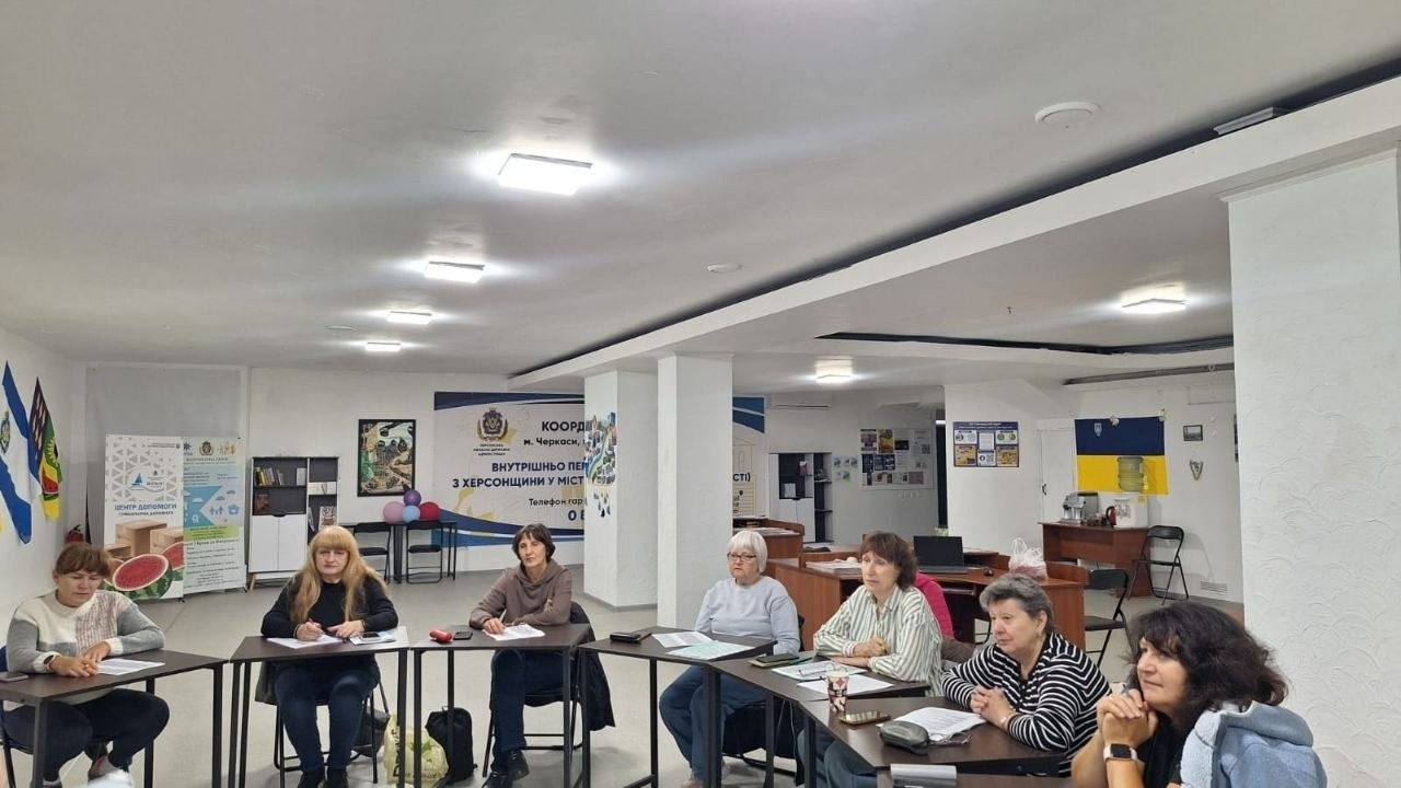 Група жінок із числа переселенців із Херсонщини сидить за розставленими півколом столами у світлому залі черкаського координаційного центру «Вільні разом» під час музичної вікторини в межах Тижня української музики. Сім учасниць заходу, організованого за сприяння Чаплинської СВА та Долматівської СВА, уважно слухають ведучих та роблять нотатки, згадуючи класику естради, сучасних виконавців та рідні пісні, що асоціюються з домом. Зустріч у межах традиційних вечорниць проходить на тлі великого офіційного банера координаційного центру допомоги ВПО з Херсонщини у місті Черкаси з гербом Херсонської ОВА, а інтер'єр просторого приміщення облаштований білими колонами, робочими столами, інформаційними стендами та закріпленим на стіні синьо-жовтим державним прапором України.