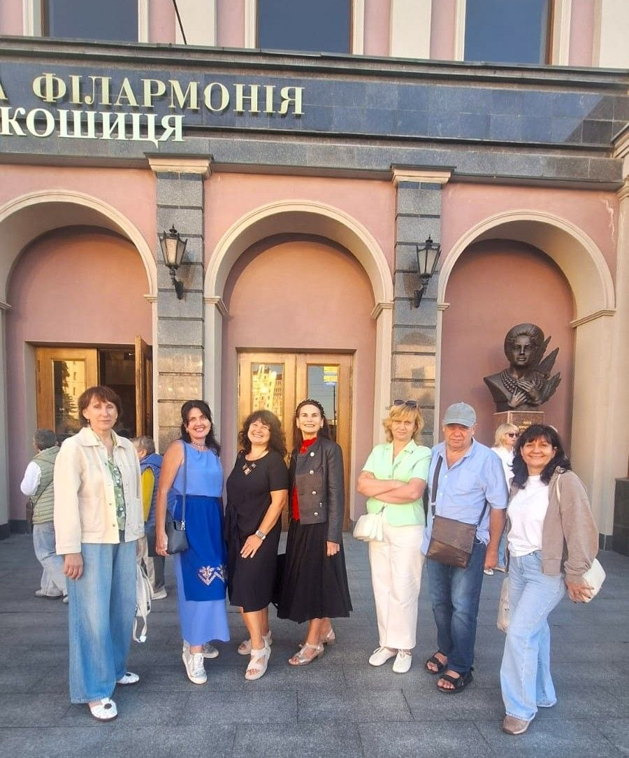 Група переселенців із Херсонщини позує перед будівлею Черкаської обласної філармонії ім. Олександра Кошиця, де вони відвідали відкриття 35-го концертного сезону Академічного симфонічного оркестру. Сім слухачів — шість жінок та один чоловік — посміхаються в камеру на тлі рожевого арочного фасаду з вивіскою «ФІЛАРМОНІЯ КОШИЦЯ» та бронзовим погруддям праворуч. Візит організовано черкаським координаційним центром «Вільні разом» за сприяння Чаплинської та Долматівської СВА в межах Тижня української музики, де гості слухали класичні твори Скорика, Бетховена та інших композиторів.