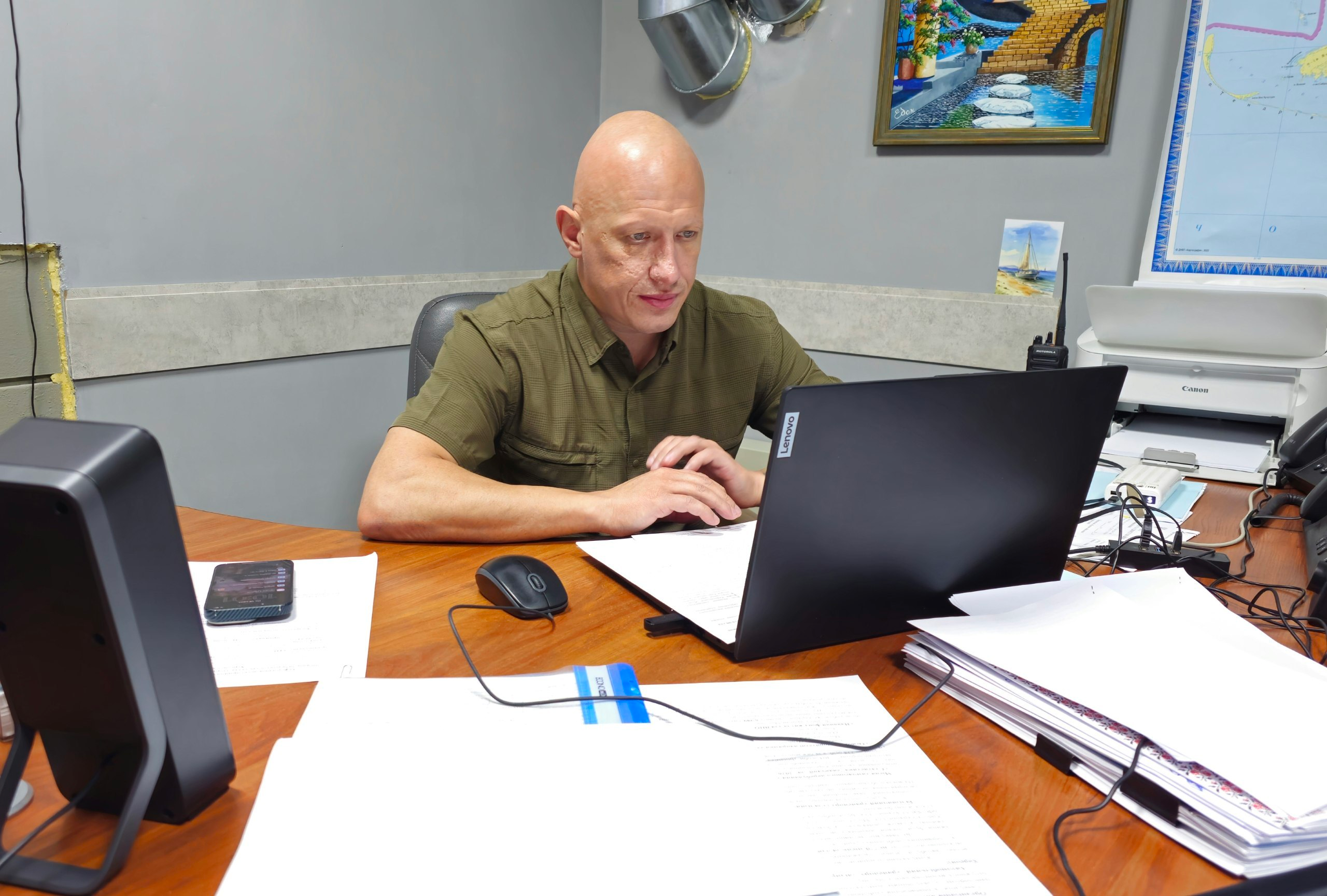 Заступник начальника Херсонської ОВА Євгеній Ігнатенко сидить за робочим столом перед відкритим ноутбуком Lenovo під час засідання комісії з питань надання одноразової матеріальної допомоги мешканцям області. Чоловік у тактичній сорочці кольору хакі зосереджено працює з документами, де розглядаються сотні звернень від громадян, які потребують фінансової підтримки внаслідок російської агресії. На столі розкладено багато робочих паперів, документів, лежить мобільний телефон, а праворуч помітно рацію та принтер. На сірій стіні позаду керівника висить картина з морським пейзажем та край великої настінної географічної карти, а за результатами засідання заявникам призначено допомогу на загальну суму 7 мільйонів 690 тисяч гривень з обласного бюджету.