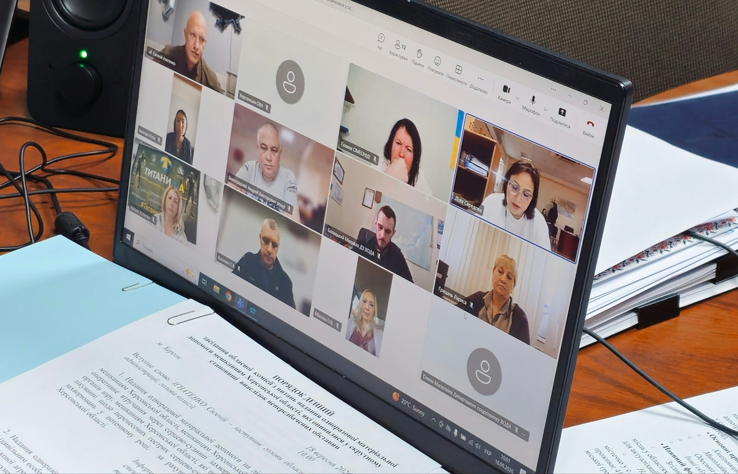 Екран ноутбука відображає онлайн-нараду в Microsoft Teams під час засідання обласної комісії з питань надання матеріальної допомоги мешканцям Херсонщини, на якому заявникам призначили загалом 7 мільйонів 690 тисяч гривень. На екрані у сітці відеодзвінка видно віконця з учасниками зустрічі, серед яких у лівому верхньому кутку відображається голова комісії, заступник начальника Херсонської ОВА Євгеній Ігнатенко, а також інші члени комісії, які розглянули 538 звернень від громадян, що потребують фінансової підтримки. Перед клавіатурою комп'ютера на столі лежать роздруковані документи, робочі папки та папери.