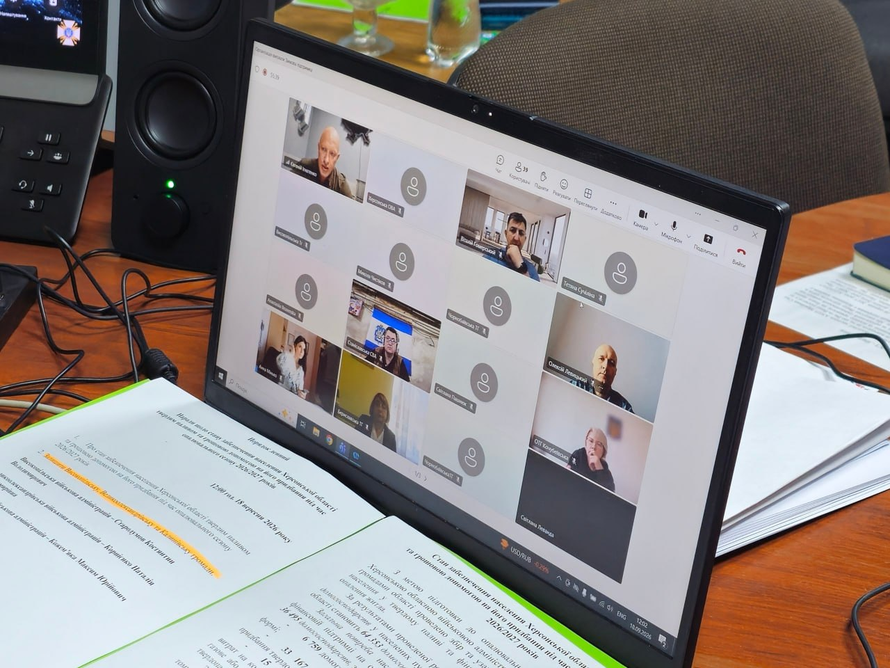 Екран ноутбука крупним планом відображає онлайн-нараду в Microsoft Teams під час засідання за участі представників деокупованих громад і підрозділів ХОДА щодо забезпечення жителів твердим паливом та грошовою допомогою. На екрані у сітці відеодзвінка видно віконця з учасниками зустрічі, серед яких у лівому верхньому кутку відображається заступник начальника Херсонської ОВА Євгеній Ігнатенко, який провів обговорення підготовки до прийдешніх холодів та опрацювання виплат за державною програмою «Зимова підтримка». Перед клавіатурою комп'ютера на столі лежать роздруковані документи, присвячені стану підготовки та допомоги населенню Херсонської області, де допомогу вже надано 837 домогосподарствам, а праворуч і ліворуч на столі розміщені інші робочі папери й папки з документами.