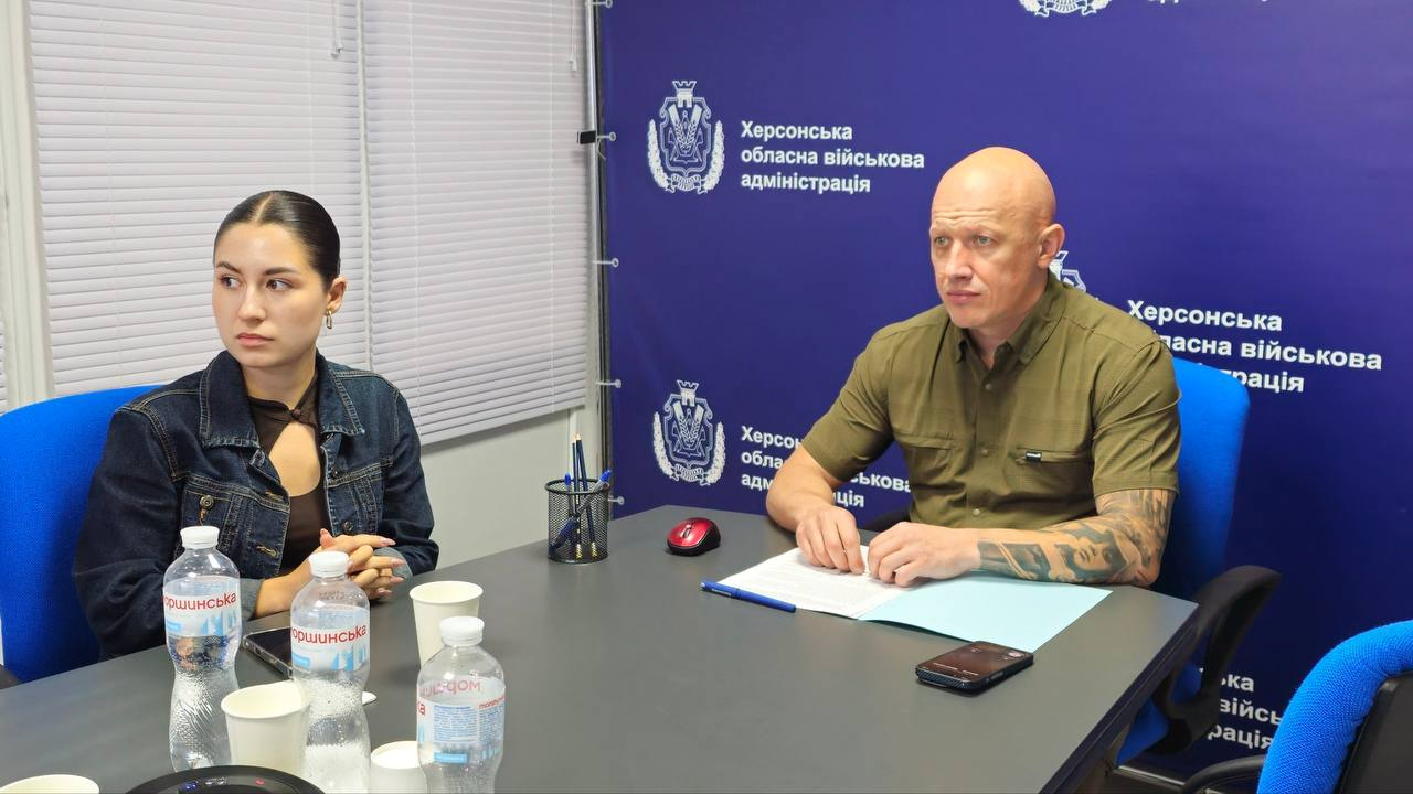Заступник начальника Херсонської ОВА Євгеній Ігнатенко та начальниця управління міжнародного співробітництва та залучення інвестицій Херсонської ОДА Марія Богодухова сидять за робочим столом під час зустрічі з організацією «People in Need» щодо підготовки громад до опалювального сезону. Євгеній Ігнатенко в тактичній сорочці кольору хакі з коротким рукавом тримає руки на столі біля робочих паперів і ручки. Ліворуч від нього розмістилася Марія Богодухова в темній джинсовій куртці, яка уважно слухає обговорення потреб прифронтових територій. Засідання проходить на тлі великого темно-синього банера з офіційними логотипами «Херсонська обласна військова адміністрація».