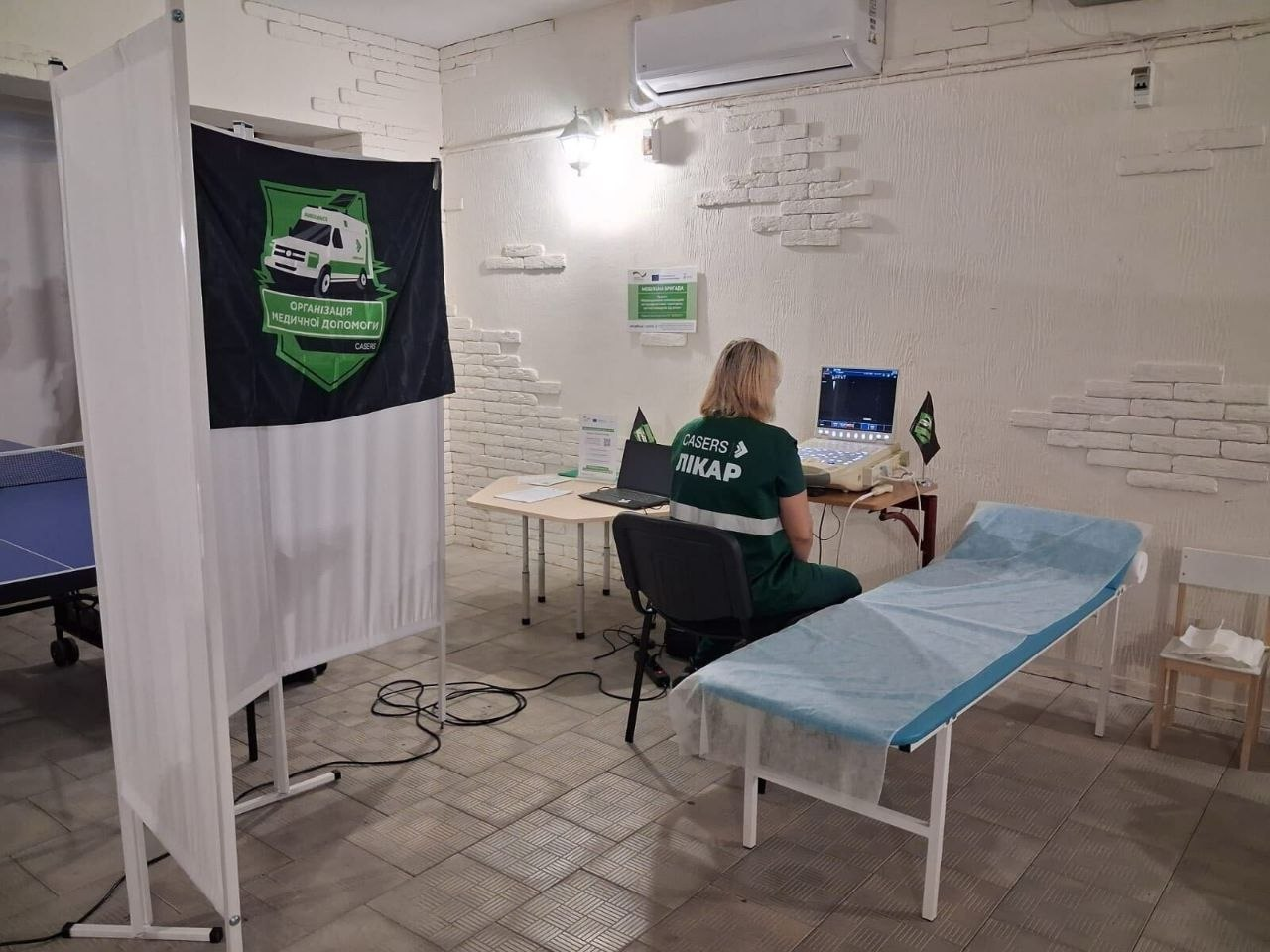 Лікарка мобільної бригади ГО «CASERS» сидить спиною до камери за робочим столом в облаштованому медичному кабінеті перед увімкненим портативним апаратом ультразвукової діагностики під час виїзду до Калинівського, Чорнобаївки та Великої Олександрівки. Жінка у темно-зеленому фірмовому жилеті з написом «ЛІКАР» налаштовує обладнання для обстеження щитоподібної залози, органів черевної порожнини та молочних залоз мешканців прифронтових громад Херсонщини. Поруч із нею розташована оглядова кушетка, накрита блакитним одноразовим простирадлом, а ліворуч стоїть додатковий стіл із ноутбуком та документами. Робоча зона організована в межах проєкту EMPOWER за підтримки GIZ Ukraine у приміщенні зі світлими стінами, де встановлено білу мобільну ширму із закріпленим на ній темним прапором організації з написом «ОРГАНІЗАЦІЯ МЕДИЧНОЇ ДОПОМОГИ CASERS», а на задньому плані помітно край тенісного столу.