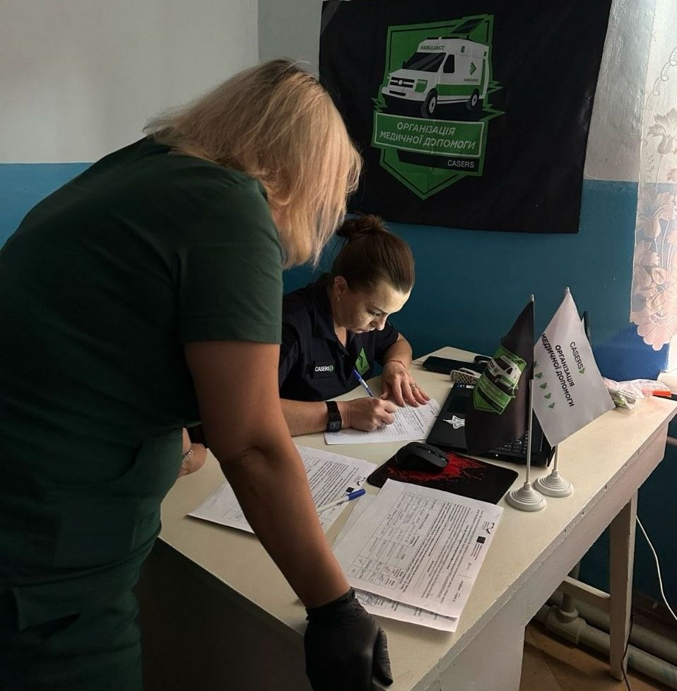 Медична працівниця у фірмовій сорочці з логотипом організації «CASERS» зосереджено заповнює документи за робочим столом під час прийому мешканців прифронтових громад у межах проєкту EMPOWER. Поруч із нею ліворуч стоїть медикиня у темно-зеленій формі та рукавичках, яка схилилася над столом і спостерігає за процесом реєстрації пацієнтів із Калинівського, Чорнобаївки та Великої Олександрівки для проведення УЗД-обстежень. На робочій поверхні столу розкладено бланки, медичні анкети, папери, лежить кулькова ручка, комп'ютерна мишка, а праворуч встановлено два настільні прапорці з фірмовою символікою ГО «CASERS». Прийом та реєстрація громадян для надання безоплатної допомоги за підтримки GIZ Ukraine проходять у кімнаті, де на задньому плані над столом висить великий темний прапор із написом «ОРГАНІЗАЦІЯ МЕДИЧНОЇ ДОПОМОГИ CASERS» та зображенням автомобіля швидкої допомоги.