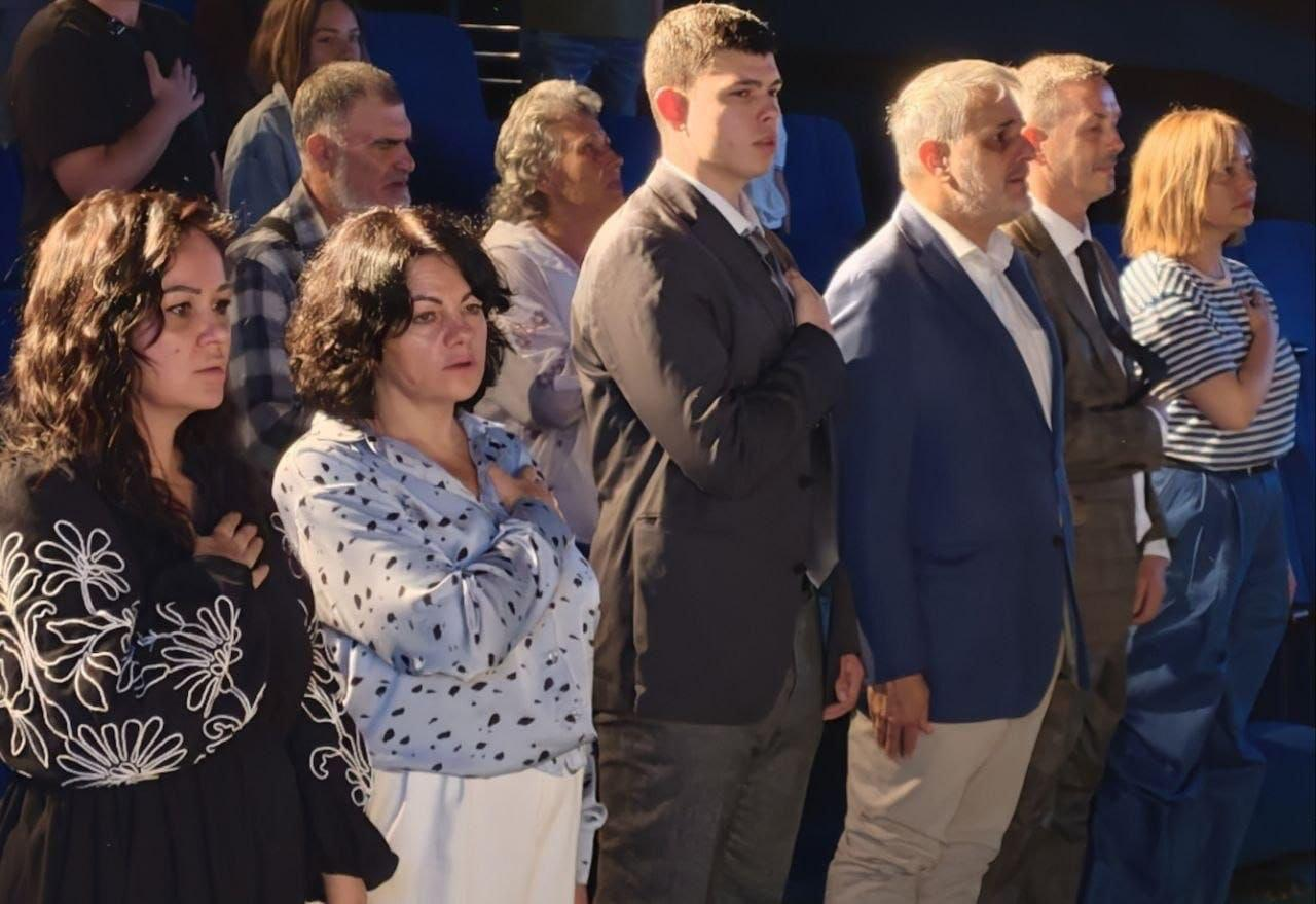 Група людей урочисто стоїть у залі, приклавши праву руку до серця під час хвилини мовчання в межах мистецького заходу «Пам’ять продовжується в музиці», присвяченого Дню української музики у Херсонській обласній філармонії. На передньому плані вишикувалися офіційні особи, слухачі та представники культури, серед яких жінка у смугастій кофті, чоловіки в ділових костюмах, молодий чоловік у темному піджаку, а ліворуч від них — директорка Департаменту реалізації гуманітарної політики Херсонської ОДА Віолетта Царькова у білій блузі в дрібний темний горошок, яка стоїть праворуч від жінки у чорній вишиванці з великим білим рослинним орнаментом. Усі присутні повернуті боком до камери, вшановують пам’ять видатних диригентів Георгія Вазіна, Олександра Дреля, Владислава Бєлявського та Юрія Керпатенка, чиє життя було пов'язане з оркестром «Гілея», на тлі синіх крісел глядацького залу.