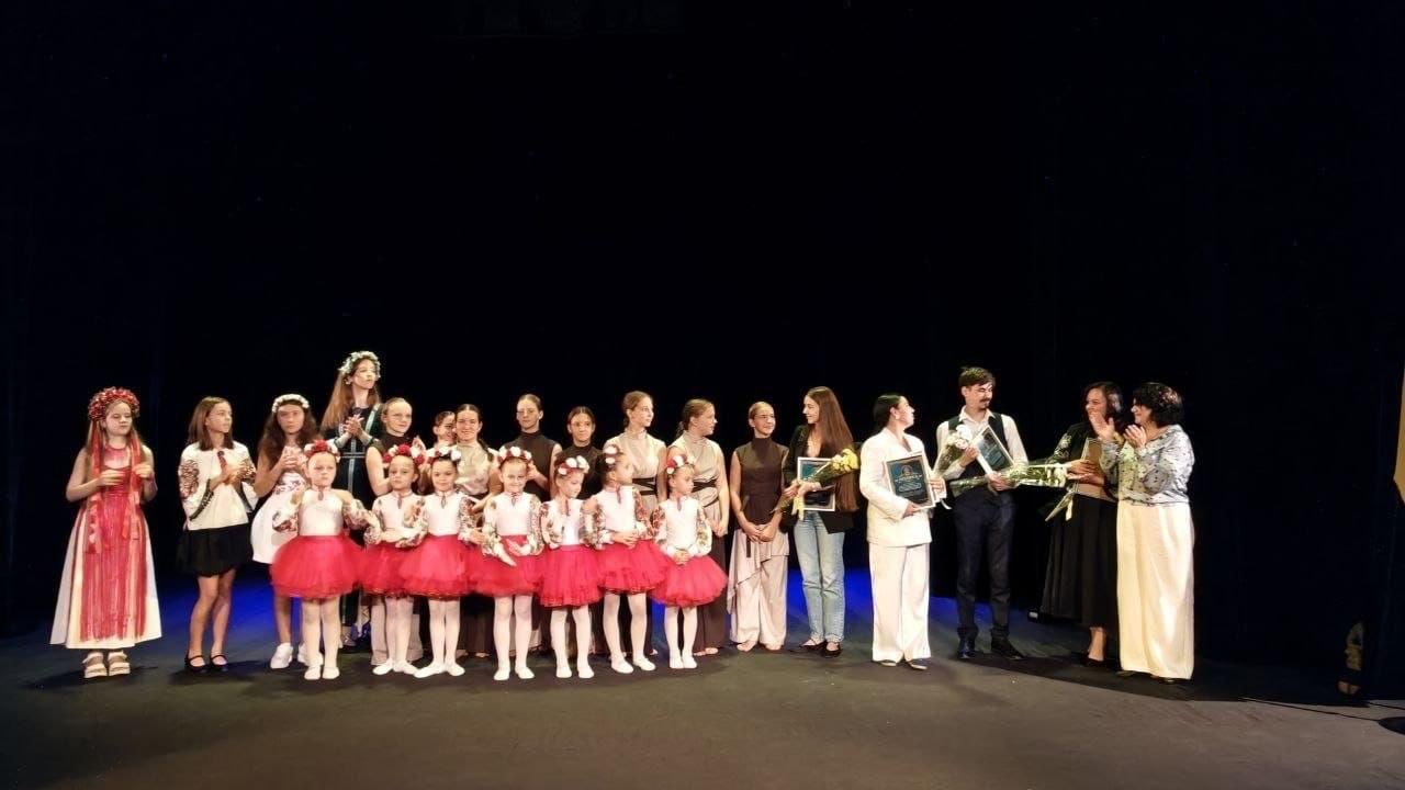 Велика група артистів та організаторів різного віку урочисто стоїть у ряд на сцені Херсонської обласної філармонії під час фіналу мистецького заходу «Пам’ять продовжується в музиці», присвяченого Дню української музики. На передньому плані ліворуч вишикувалися юні учасниці дитячого колективу в пишних червоних спідницях-пачках та білих вишиванках, а також дівчатка у традиційних українських віночках із довгими стрічками та вишитих сукнях. Праворуч розмістилися дорослі солісти та представники творчих колективів філармонії в сценічних костюмах і сукнях, які тримають у руках букети квітів, нагородні грамоти у рамках та аплодують глядачам після завершення концертної програми, що була присвячена пам'яті видатних диригентів Георгія Вазіна, Олександра Дреля, Владислава Бєлявського та Юрія Керпатенка. Усі присутні повернуті обличчям до залу на великій сцені на тлі глибокої чорної куліси.