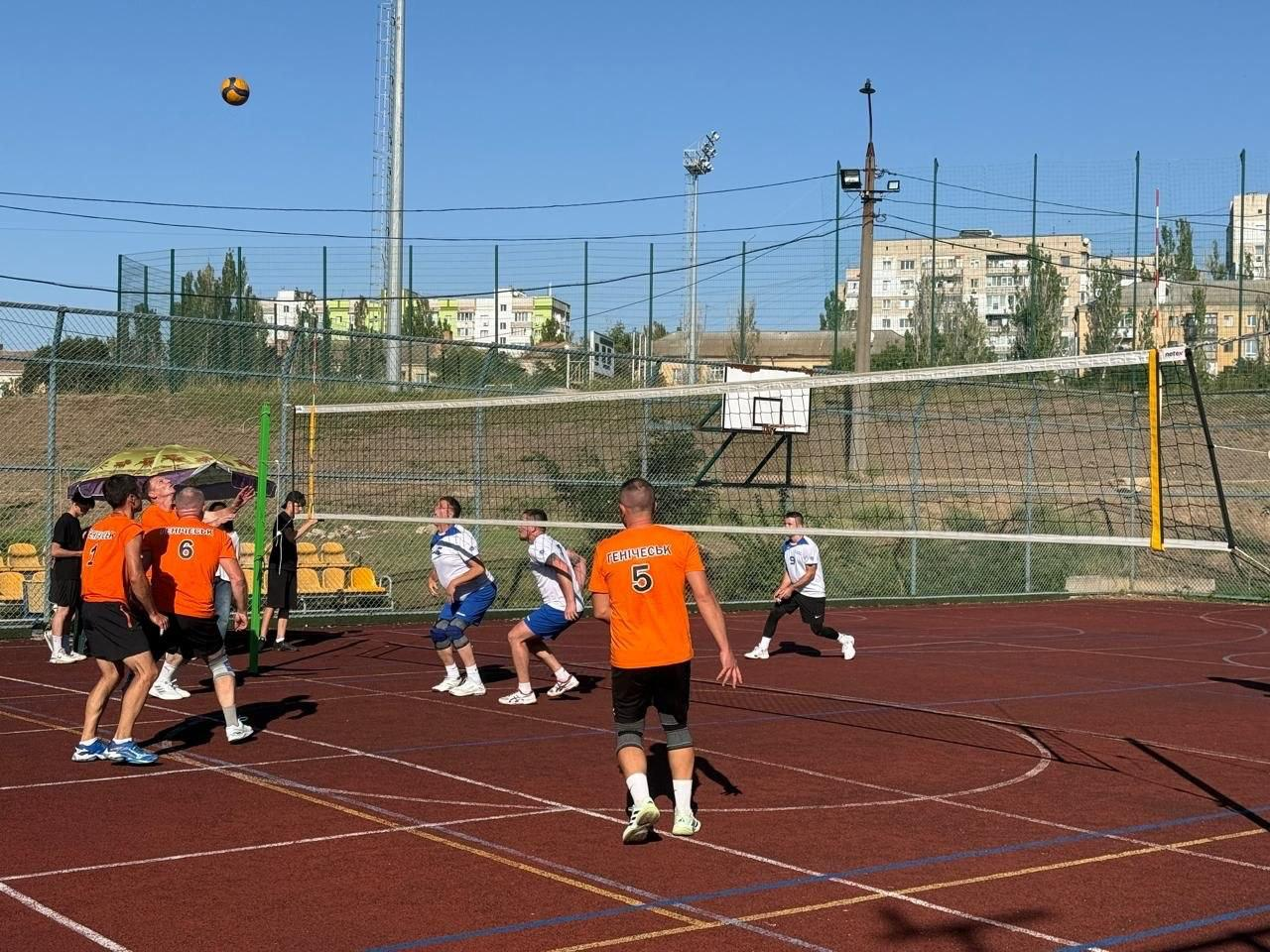 Ігровий момент волейбольного матчу під час відкритого Херсонського обласного турніру з паркового волейболу серед чоловічих команд, присвяченого Дню спорту та річницям заснування Херсона й Генічеська. На передньому плані майданчика з червоно-коричневим гумовим покриттям спиною до камери стоїть гравець під номером 5 із написом «ГЕНІЧЕСЬК» на помаранчевій футболці, а ліворуч біля сітки згрупувалися ще троє його партнерів по команді, які представляють нині окуповану рідну громаду. З іншого боку волейбольної сітки гравці суперника у біло-блакитній формі зосереджено стежать за траєкторією польоту м'яча, що летить у небі, намагаючись захистити свій бік поля. Змагання шести колективів проходять у сонячний день на тлі металевої огорожі, глядацьких трибун та житлових будинків, де за підсумками ігор команда Генічеська виборола почесне друге місце, поступившись волейбольному клубу «Бабай» із Чорнобаївки та випередивши «Молодь Херсонщини».