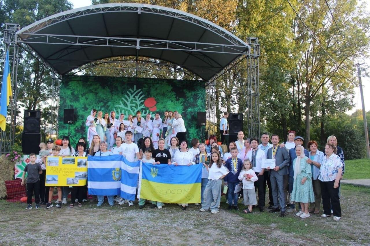 Велика група артистів, організаторів та учасників різного віку урочисто зібралася біля відкритої сцени у Великому Кучурові на Буковині після завершення VIII Всеукраїнського фестивалю «Україна барвінкова», присвяченого Дню міста-героя Херсона. На самій сцені під куполоподібним навісом вишикувалися юні учасниці творчого колективу в білих українських вишиванках. Перед сценою стоять діти й дорослі з Херсонщини та Буковини, які тримають великий синьо-жовтий Прапор України з написом «Херсон» та намальованим серденьком, біло-синій прапор міста Херсон із гербом та жовтий інформаційний плакат. Праворуч у першому ряду розмістилися представники Великокучурівської громади, організатори з Департаменту реалізації гуманітарної політики ХОДА та Херсонського фахового коледжу культури і мистецтв, які тримають у руках офіційні нагородні дипломи в рамках на тлі високих зелених дерев у сонячний день.