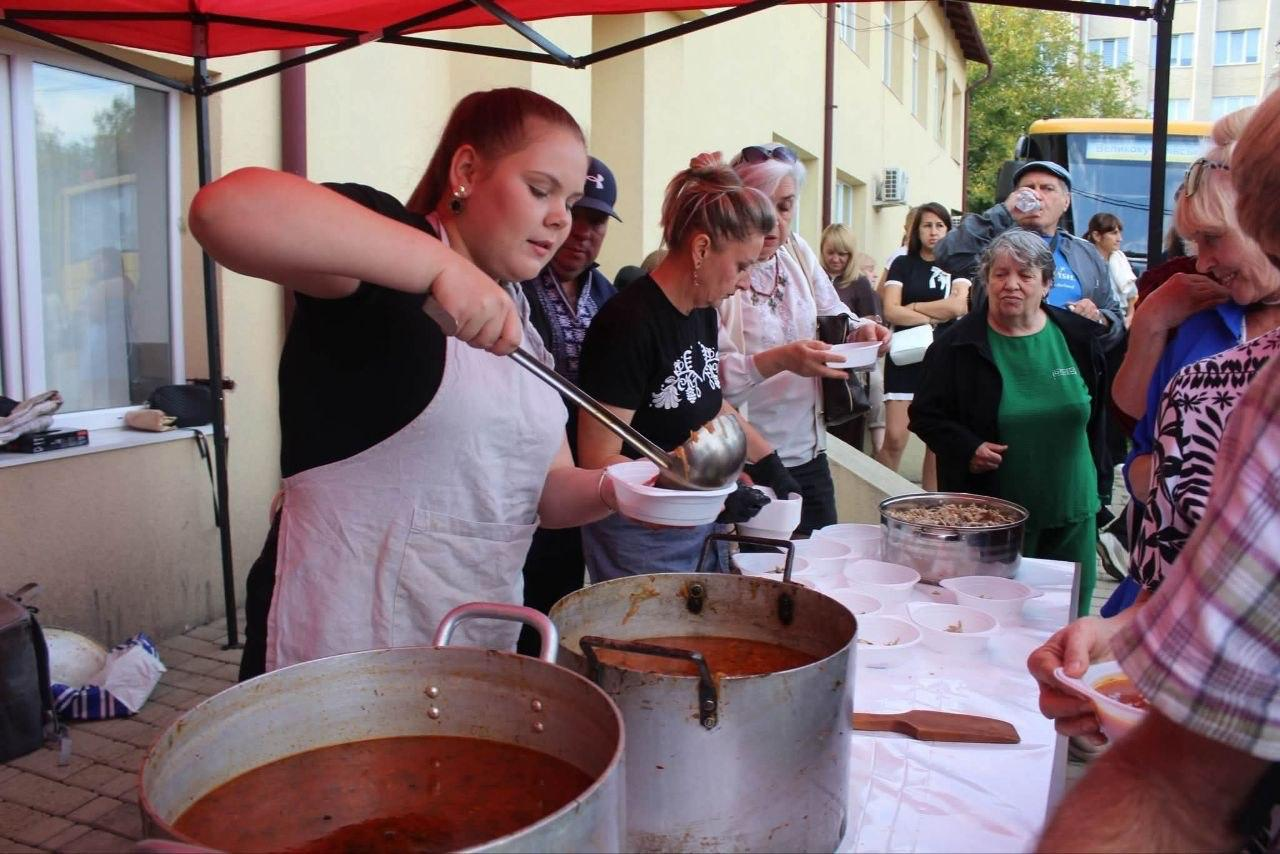 На майданчику просто неба під червоним навісом організатори розливають херсонський борщ та традиційні буковинські страви з великих алюмінієвих каструль під час VIII Всеукраїнського фестивалю «Україна барвінкова» у Великому Кучурові. Ліворуч на передньому плані молода дівчина у світлому фартуху великим ополоником наповнює глибоку білу одноразову тарілку гарячим борщем для численних гостей, які вишикувалися в довгу чергу на подвір'ї. Поруч із нею інша представниця організаторів у чорній футболці з національним орнаментом також розкладає частування в порційні тарілки, розставлені на довгому столі з білою скатертиною, де в межах свята до Дня міста-героя Херсона дорослі та діти з творчого табору можуть безкоштовно скуштувати страви херсонської та буковинської кухні на тлі світлої будівлі.