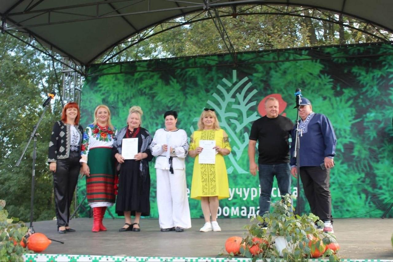 На великій відкритій сцені під куполоподібним навісом у ряд вишикувалися організатори та почесні гості фестивалю, тримаючи в руках офіційні дипломи та мікрофон. У лівій частині сцени стоїть рудоволоса жінка у темному вишитому жакеті, поруч із якою розмістилася учасниця в яскравому національному костюмі з великим квітковим вінком та червоних чоботях. По центру стоять дві жінки у довгих сукнях (чорній та яскраво-жовтій) з нагородними грамотами в руках, а між ними — ведуча чи спікерка у білому вбранні з мікрофоном. Праворуч композицію завершують двоє чоловіків: один у чорній футболці та джинсах, а інший — у синій вишиванці та синій кепці. Виступ проходить на тлі великого зеленого банера з логотипом Великокучурівської громади, а передній край сцени прикрашений композиціями із зеленого листя та помаранчевих гарбузів.