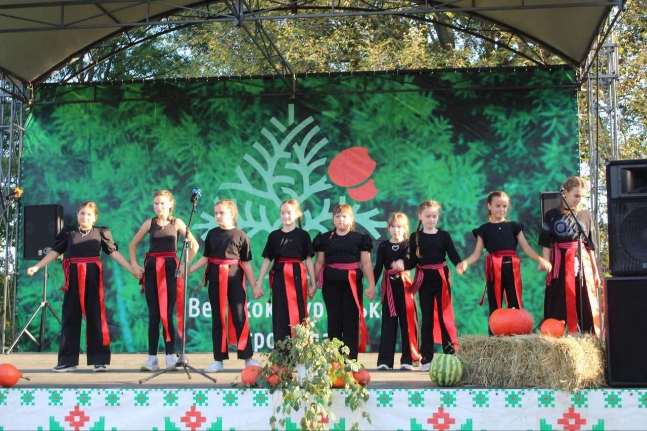 Хореографічний колектив із дев'яти дівчаток у чорних сценічних костюмах із яскраво-червоними поясами вишикувався в одну шеренгу, тримаючись за руки, на великій відкритій сцені у Великому Кучурові на Буковині під час VIII Всеукраїнського фестивалю «Україна барвінкова». Юні вихованки третього творчого табору для дітей Херсонщини стоять обличчям до глядачів перед мікрофонною стійкою, представляючи свій рідний край далеко від дому на заході, присвяченому Дню української музики та Дню міста-героя Херсона. Виступ проходить на тлі великого зеленого банера Великокучурівської громади, а край сцени прикрашений святковим декором із помаранчевих гарбузів, снопа сіна та стиглого кавуна, яким пригощали гостей свята.