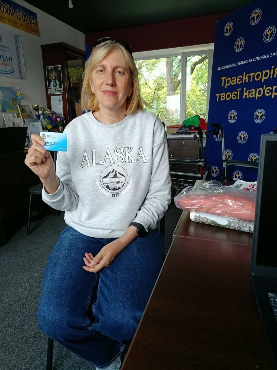 Жінка у сірому світшоті з написом «ALASKA» та синіх джинсах сидить у кабінеті й тримає в руці блакитну пластикову картку, отриману в межах реалізації проєкту «Картка Херсонщини». Вона посміхається, демонструючи картку від Координаційного центру «Вільні разом», який в Івано-Франківську здійснює комплексну підтримку переселенців. Праворуч на задньому плані встановлено великий синій банер Херсонської обласної служби зайнятості з гаслом «Траєкторія твоєї кар'єри», на передньому плані на столі лежать запаковані речі та текстильний виріб з українською вишивкою для ВПО, а ліворуч видніється офісна шафа біля великого вікна.