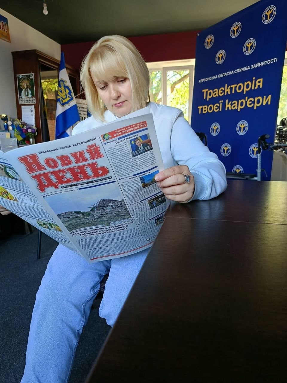 Жінка у світлій кофті та джинсах сидить за темно-коричневим столом в офісі та зосереджено читає розгорнуту друковану газету «Новий день», примірник якої переселенці отримали в межах комплексної підтримки від Координаційного центру «Вільні разом». Вона схилила голову над першою шпальтою незалежного обласного видання Херсонщини, де помітно великий червоний заголовок газети та статті про життя мешканців деокупованих територій на тлі великого синього банера Херсонської обласної служби зайнятості з гаслом «Траєкторія твоєї кар'єри». Ліворуч на задньому плані біля вікна з видом на зелені дерева видніється кабінетна шафа та Державний Прапор України, що підкреслює діяльність хабу з допомоги ВПО в Івано-Франківську.