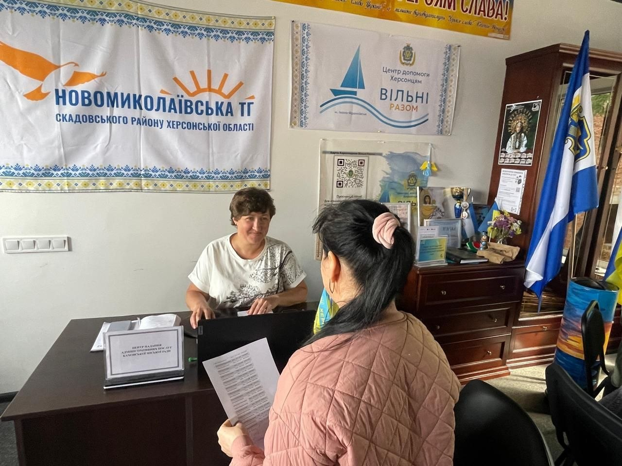 Фахівці відділів ЦНАП Каховської громади надають адміністративні послуги та консультації відвідувачам у приміщенні Центру допомоги херсонцям «Вільні разом» в Івано-Франківську. За робочим столом перед відкритим ноутбуком сидить співробітниця хабу у світлій футболці з малюнком, яка посміхається та спілкується з жінкою в рожевій куртці, що сидить спиною до камери та тримає в руках роздрукований документ. Консультування ВПО з Херсонщини проходить у кабінеті, де на стіні за ними закріплено великий білий прапор із написом «НОВОМИКОЛАЇВСЬКА ТГ СКАДОВСЬКОГО РАЙОНУ ХЕРСОНСЬКОЇ ОБЛАСТІ», поруч висить емблема Центру «Вільні разом» із синім корабликом, а праворуч на шафі встановлено прапор Херсонської області.