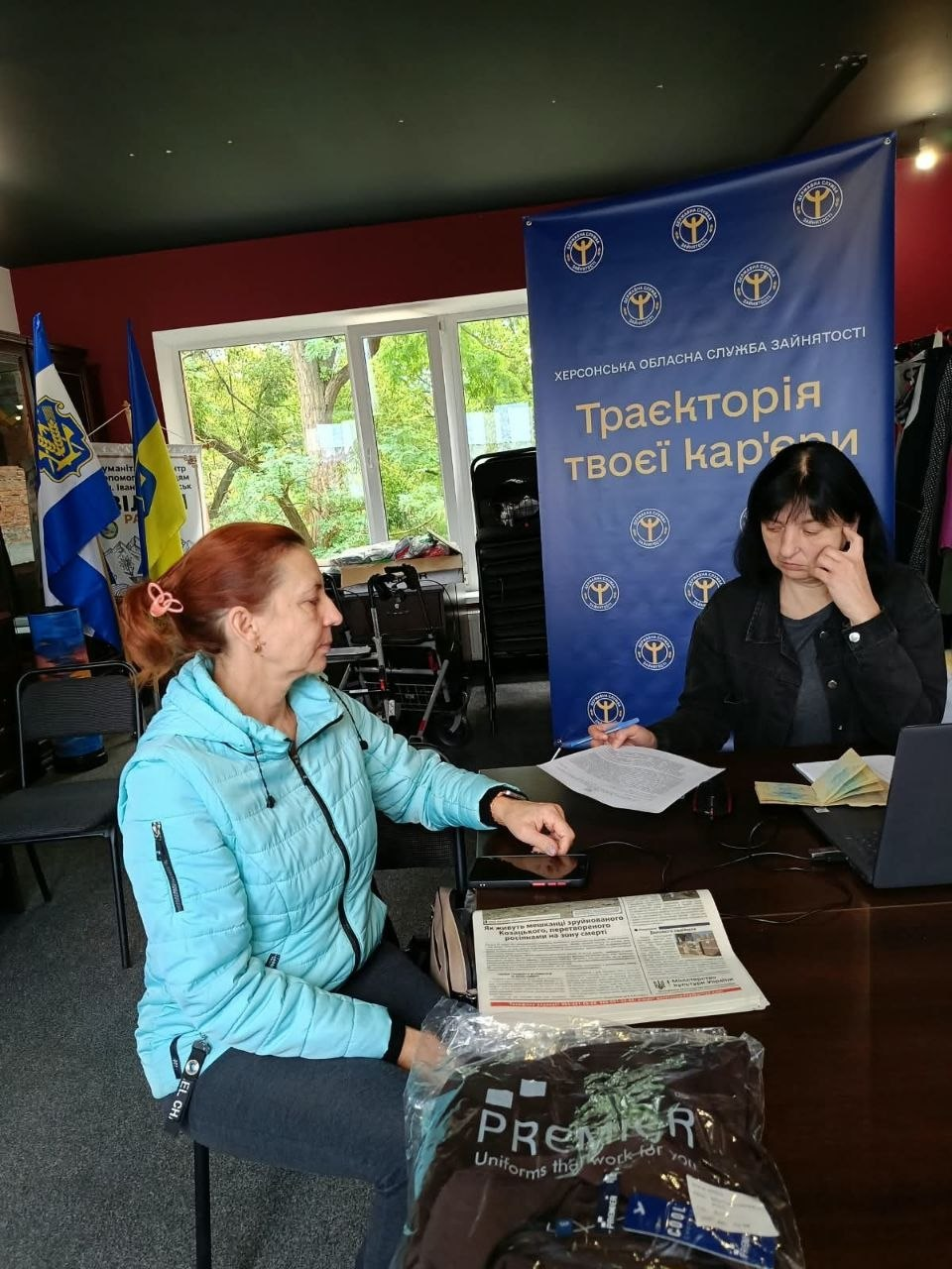 Фахівчиня Херсонського міського центру зайнятості надає консультації щодо працевлаштування відвідувачці у приміщенні Координаційного центру «Вільні разом» в Івано-Франківську. Ліворуч сидить рудоволоса жінка у блакитній куртці, яка уважно слухає спеціалістку та тримає руку на столі поруч зі своїм смартфоном. За робочим столом праворуч сидить співробітниця у темній куртці, яка переглядає роздруковані офіційні документи переселенки перед відкритим ноутбуком, де для комплексної підтримки ВПО також підготовлено друковану пресу та новий запакований одяг. Консультування громадян проходить на тлі великого брендованого синього банера Херсонської обласної служби зайнятості з гаслом «Траєкторія твоєї кар'єри», а ліворуч на задньому плані біля вікна встановлено Державний Прапор України та прапор Херсонської області.