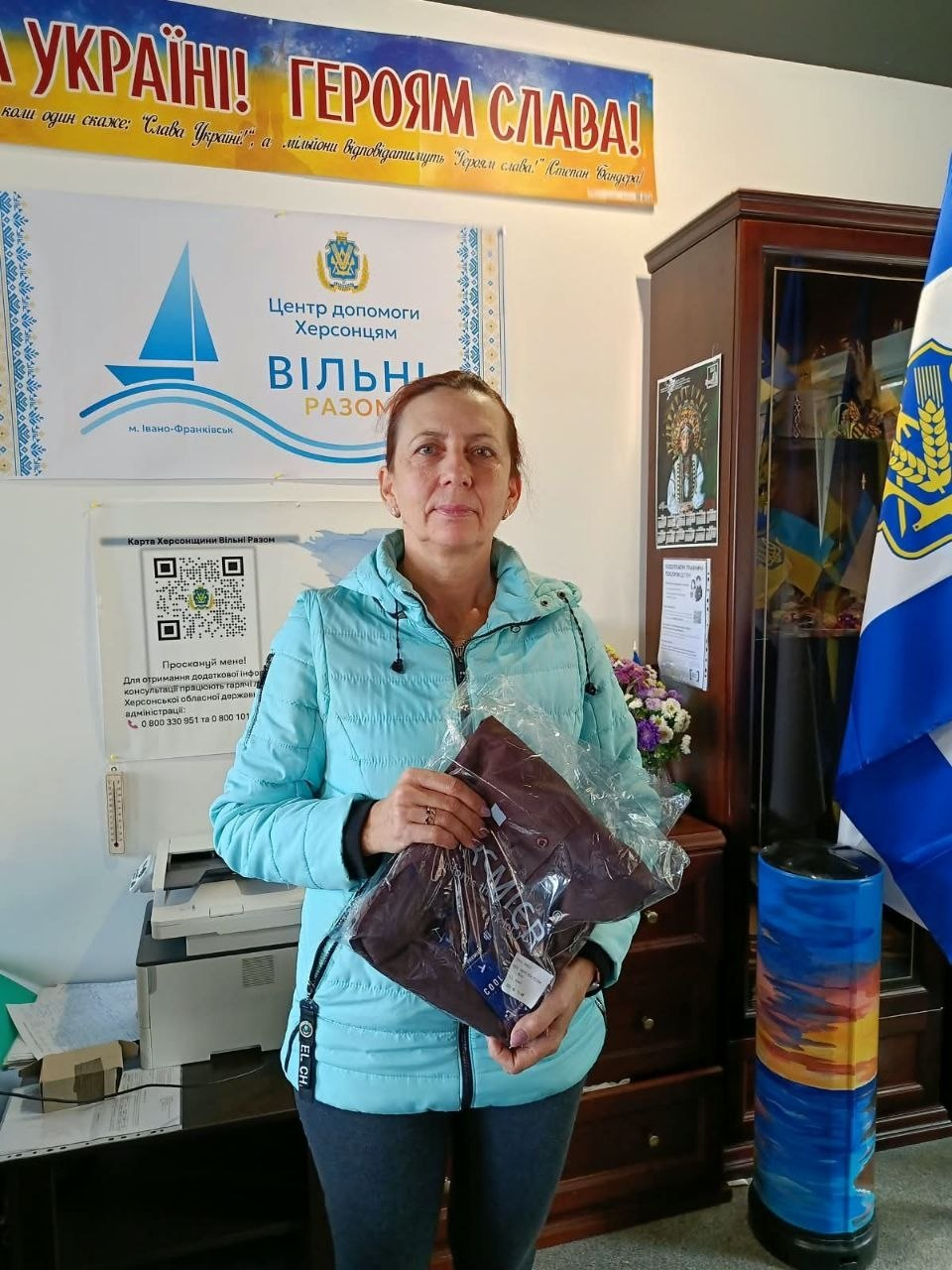 Жінка середнього віку у світло-блакитній куртці стоїть у приміщенні хабу в Івано-Франківську та тримає в руках новий сезонний одяг у прозорій поліетиленовій упаковці, виданий у межах комплексної підтримки переселенців з Херсонщини. Вона повернута обличчям до камери та тримає пакунок обома руками, отримавши гуманітарну допомогу від благодійних фондів для забезпечення ВПО. На стіні за нею ліворуч закріплено білу емблему з синім корабликом та написом «Центр допомоги херсонцям ВІЛЬНІ РАЗОМ м. Івано-Франківськ», під якою розміщено плакат із QR-кодами «Картка Херсонщини Вільні Разом». У верхній частині стіни висить патріотичний банер із гаслом «СЛАВА УКРАЇНІ! ГЕРОЯМ СЛАВА!», праворуч стоїть висока дерев'яна офісна шафа, а на передньому плані праворуч видніється край синьо-білого прапора Херсонської області.