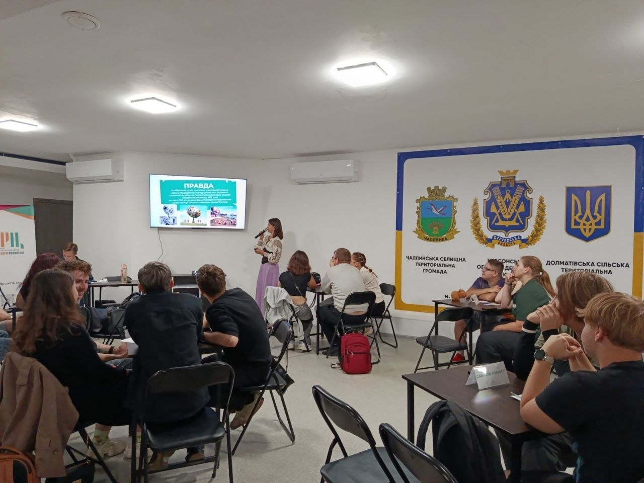 Учасники інтелектуально-розважальної вікторини «Відкривай для себе Україну» сидять групами за столами у залі Координаційного центру «Вільні разом» у Черкасах, де для молоді віком від 14 до 35 років організували інтерактивний захід. Хлопці та дівчата з числа переселенців із Херсонщини зосереджено слухають спікерку з мікрофоном, яка стоїть біля настінного екрана з презентаційним слайдом «ПРАВДА» у межах просвітницької програми, створеної у співпраці з Черкаським молодіжним ресурсним центром. На правій стіні світлої кімнати розміщено великий інформаційний банер із символікою та офіційними назвами Чаплинської селищної, Херсонської обласної та Долматівської сільської територіальних громад, а самі учасники змагання активно обговорюють завдання турніру в командах.