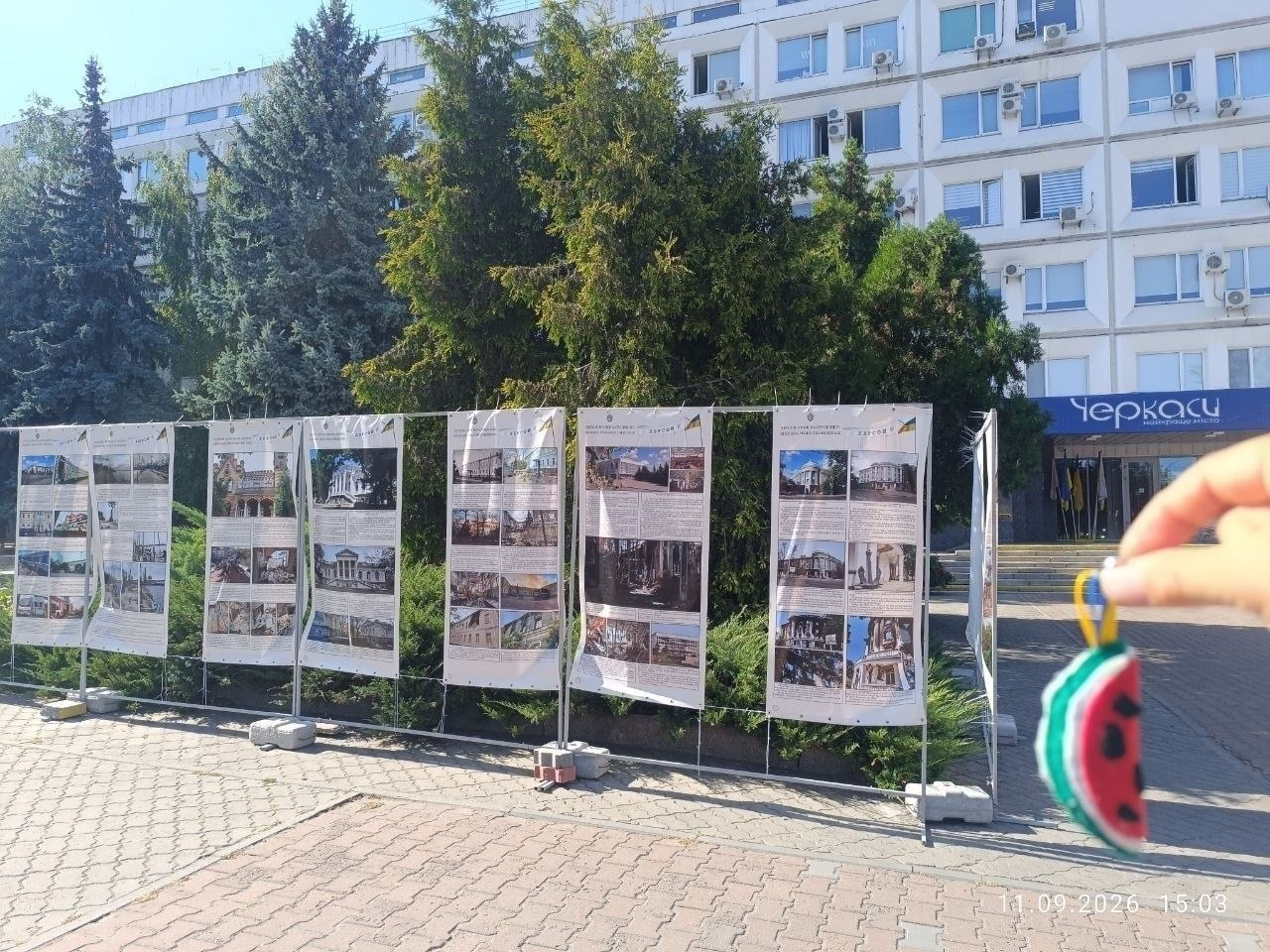На площі перед будівлею міської ради Черкас розгорнуто велику вуличну фотовиставку «Херсон. Зранена спадщина», присвячену пошкодженим пам'яткам архітектури, яка відкрилася в межах культурних заходів та всебічної підтримки переселенців завдяки Координаційному центру «Вільні разом». Стенди містять численні світлини історичних споруд і культурних об'єктів Херсонщини, що зазнали руйнувань, а праворуч на передньому плані в руці людини зафіксовано в'язаний червоно-зелений сувенірний брелок у вигляді скибочки кавуна. На задньому плані височіє багатоповерхова світла адміністративна будівля, над входом до якої закріплено синій напис «Черкаси — місто вільних людей», а самі інформаційні плакати встановлені на тротуарній плитці вздовж густих зелених дерев у сонячний день.