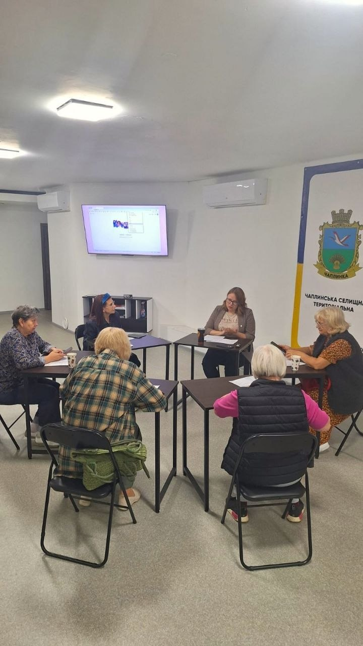 Група жінок сидить колом за невеликими столами у світлому приміщенні Координаційного центру «Вільні разом» у Черкасах під час регулярного заняття з англійської мови, де для всебічної підтримки переселенців з Херсонщини проводиться практичне навчання. У центрі кімнати за двома столами сидять дві викладачки, які ведуть бесіду з відвідувачками та допомагають їм розбирати композиції з класичних мюзиклів «Cats» та «Funny Girl». Ліворуч на стіні встановлено телевізійний екран, на якому відображається робочий інтерфейс програми з двома відкритими вікнами на світлому тлі, що слугує наочним матеріалом для вивчення іноземної мови. Праворуч на стіні закріплено великий білий банер із гербом селища Чаплинка та офіційним написом Чаплинська селищна територіальна громада, а самі слухачки розмістилися на розкладних стільцях під час обговорення навчального матеріалу.