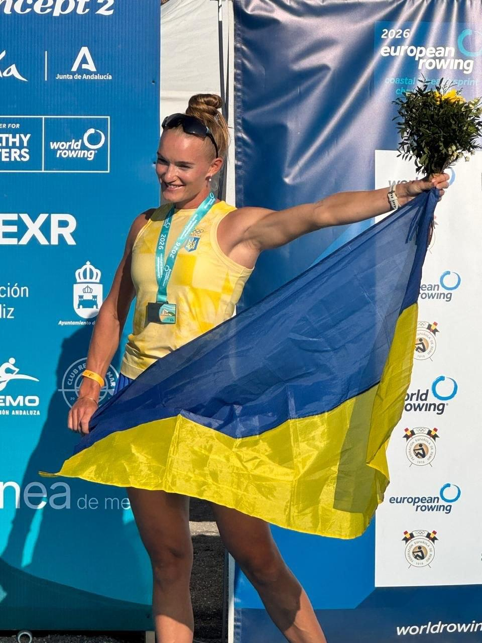 Заслужений майстер спорту України зі Скадовська Катерина Дудченко урочисто позує на п'єдесталі пошани чемпіонату Європи з пляжного прибережного веслування в Іспанії, де вона виборола «срібло» в одиночці та «золото» в жіночій двійці, розгорнувши за спиною синьо-жовтий Державний Прапор України. Спортсменка Херсонської школи вищої спортивної майстерності у жовтому топі з національним гербом широко посміхається та піднімає вгору ліву руку із жовтим святковим букетом квітів, демонструючи дві здобуті медалі на бірюзових стрічках за результатами триденних змагань у Ла-Лінеа-де-ла-Консепсьйон. Вона зафіксована великим планом на тлі офіційного брендованого банера турніру «2026 European Rowing Coastal & Beach Sprint Championships», де нанесені логотипи міжнародних організацій «world rowing», «Concept 2» та емблеми спонсорів.