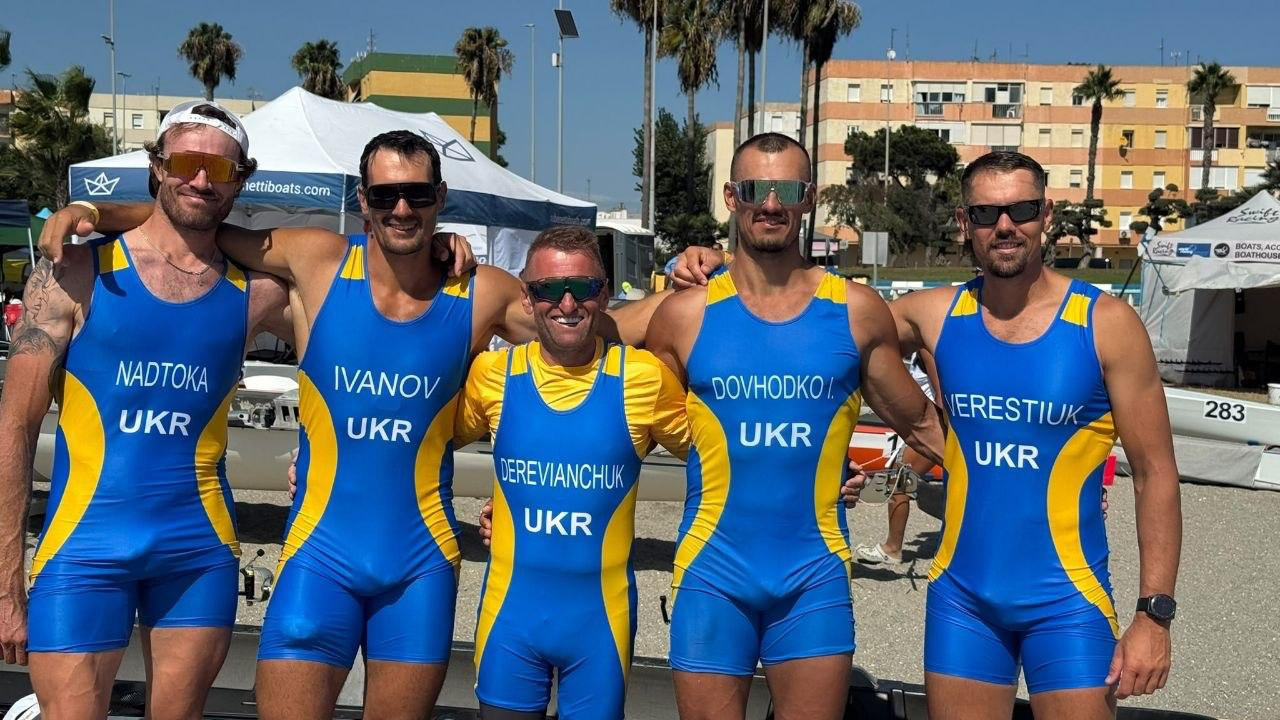 Чоловічий екіпаж української четвірки зі стерновим згуртовано позує в обнімку на морському узбережжі Іспанії після виборення срібної нагороди 20 вересня на чемпіонаті Європи з пляжного прибережного веслування у Ла-Лінеа-де-ла-Консепсьйон. П'ятеро спортсменів стоять у ряд, широко посміхаючись у сонцезахисних окулярах, вдягнені в однакові синьо-жовті облягаючі спортивні комбінезони з написами «UKR» та прізвищами на грудях. Зліва направо вишикувалися: Олександр Надтока («NADTOKA»), Юрій Іванов («IVANOV»), головний тренер збірної України та стерновий Андрій Дерев’янчук («DEREVIANCHUK»), Іван Довгодко («DOVHODKO I.»), а також майстер спорту України міжнародного класу, спортсмен Херсонської школи вищої спортивної майстерності Артем Верестюк («VERESTIUK») праворуч. Вони сфотографовані просто неба на тлі білих наметів, веслувальних човнів, високих пальм та багатоповерхових житлових будинків під яскравим сонцем за результатами триденного турніру.
