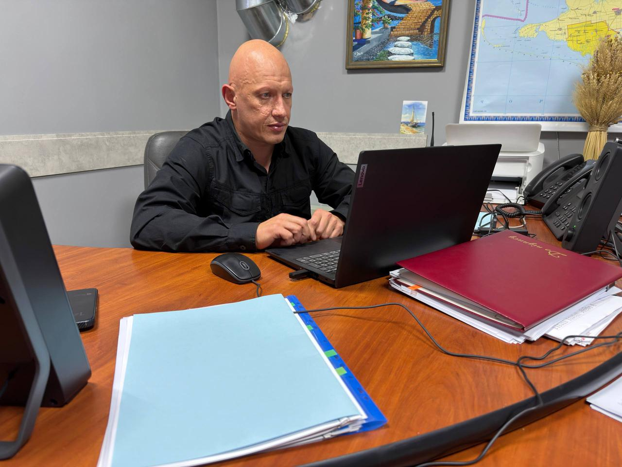 Заступник начальника Херсонської ОВА Євгеній Ігнатенко за робочим столом перед відкритим ноутбуком проводить онлайн-зустріч із представниками громад щодо покращення ефективності та злагодженості роботи 14 гуманітарних центрів «Вільні разом», які допомагають переселенцям з Херсонщини по всій Україні. Посадовець у чорній сорочці сидить у кабінеті, де на передньому плані на великому дерев'яному столі лежать робочі теки, зокрема бордова папка з документами «До підпису». На задньому плані на сірій стіні висить картина з морським краєвидом, а праворуч закріплено карту України, біля якої встановлено офісну техніку, стаціонарний телефон та декоративний сніп пшениці, що ілюструє проведення наради з обговорення поточної діяльності, проблемних питань та розвитку осередків підтримки ВПО.