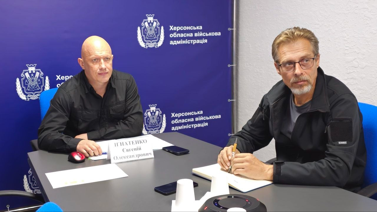 Заступник начальника Херсонської ОВА Євгеній Ігнатенко та начальник управління планування заходів цивільного захисту та матеріального забезпечення ХОВА Володимир Бєляєв сидять поруч за робочим столом під час координаційної наради під головуванням Міністра у справах громад, територій та внутрішньо переміщених осіб України Віталія Безгіна. Ліворуч за столом сидить чоловік у чорній тактичній сорочці — Євгеній Ігнатенко, перед яким стоїть біла настільна табличка з його ім'ям, а праворуч розмістився чоловік в окулярах у чорній куртці — Володимир Бєляєв, який тримає в руці ручку над відкритим блокнотом. Посадовці зафіксовані на тлі великого синього брендованого банера з офіційними гербами та повторюваними білими написями «Херсонська обласна військова адміністрація» під час обговорення урядових доручень щодо життєдіяльності та комплексної безпеки прифронтових регіонів, а також пропозицій від Херсонської ОВА стосовно розширення вмісту «Пакунку тепла» для підтримки цивільного населення деокупованих територій перед початком холодів.
