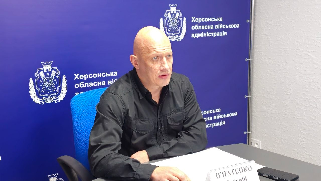 Заступник начальника Херсонської ОВА Євгеній Ігнатенко сидить за робочим столом під час координаційної наради під головуванням Міністра у справах громад, територій та внутрішньо переміщених осіб України Віталія Безгіна щодо забезпечення життєдіяльності прифронтових регіонів. Посадовець у чорній тактичній сорочці зафіксований у синьому офісному кріслі, спершись руками на стіл, де перед ним лежать документи та встановлено білу табличку з його прізвищем. Кадр зроблено на тлі синього брендованого банера з гербами та повторюваними написями «Херсонська обласна військова адміністрація» під час обговорення пропозицій щодо розширення вмісту «Пакунку тепла» для понад 111 тисяч жителів пільгових категорій деокупованої Херсонщини, що включатиме засоби автономного живлення, медикаменти та газові обігрівачі перед початком холодів.
