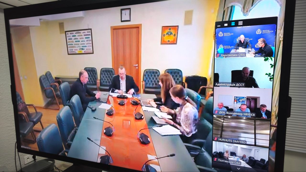 Великий телевізійний екран відображає координаційну онлайн-нараду під головуванням Міністра у справах громад, територій та внутрішньо переміщених осіб України Віталія Безгіна щодо комплексної безпеки прифронтових регіонів. На лівій частині екрана зафіксовано залу засідань міністерства, де за столом із мікрофонами працюють четверо осіб, а на правій бічній панелі виведено вікна інших підключених учасників конференції. У верхньому віконці з підписом «Херсонська ОВА» видно заступника начальника ХОВА Євгенія Ігнатенка та начальника управління Володимира Бєляєва під час обговорення пропозицій щодо розширення «Пакунку тепла» для жителів деокупованої Херсонщини, а нижче відображаються вікна «Адміністрація ДССТ», «ДКА, Володимир МІХЄЄВ», «HURHUTS DMYTRO» та «Полтавська ОВА», що ілюструє спільну роботу з Урядом задля безпекових та гуманітарних потреб області.