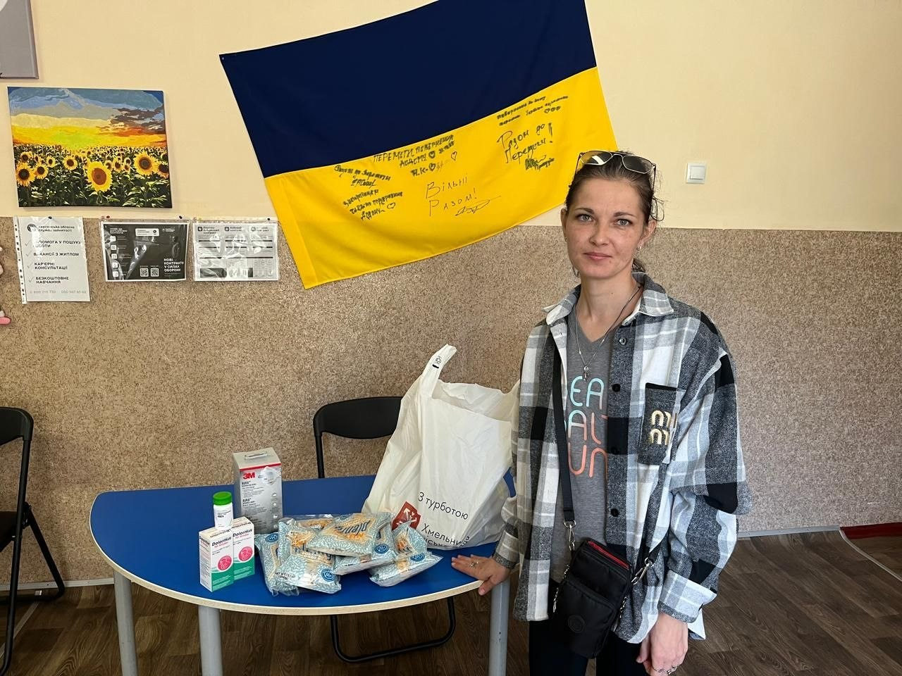 Жінка у картатій чорно-білій сорочці стоїть у приміщенні біля синього напівкруглого столу, на якому розкладено медичні засоби та пакунки з гуманітарною допомогою в межах системної підтримки ВПО з Херсонщини від Координаційного центру «Вільні разом» у Хмельницькому. Вона дивиться у камеру, поклавши руку на великий білий пакет із написом «З турботою... Хмельницький...», поруч із яким для видачі переселенцям підготовлено лікарські засоби, коробку із захисними масками «3M», одноразові медичні халати та шприци. На задньому плані на світло-бежевій стіні закріплено великий синьо-жовтий Державний Прапор України з патріотичними написами та побажаннями від руки, а ліворуч висять інформаційні листки та картина із соняшниковим полем, що ілюструє передачу тридцяти семи одиниць ліків, вісімдесяти масок, двадцяти халатів та сімнадцяти шприців.