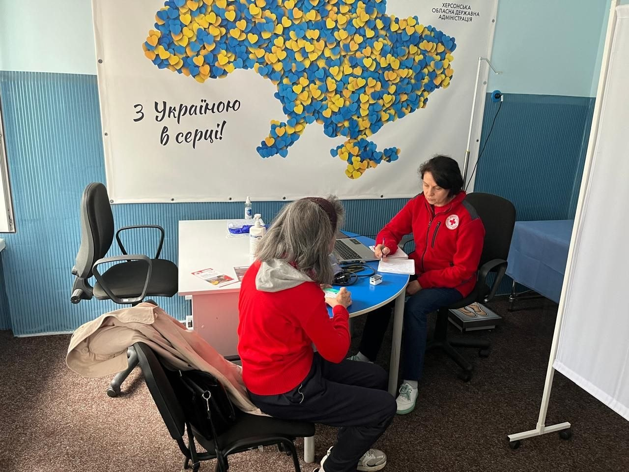 Медична працівниця у червоній куртці з емблемою Червоного Хреста та пацієнтка у червоній кофті сидять за робочим столом у кабінеті під час виїзного прийому мобільної бригади Товариства Червоного Хреста України, яка організована для надання медичної допомоги в межах системної підтримки ВПО з Херсонщини від Координаційного центру «Вільні разом» у Хмельницькому. Лікарка робить записи ручкою на папері перед відкритим ноутбуком, консультуючи відвідувачку, яка сидить спиною до камери біля тонометра та медичних буклетів, де переселенці можуть безкоштовно отримати консультації та необхідні медикаменти. На задньому плані на стіні висить великий білий банер із патріотичним написом «З Україною в серці!», мапою України з маленьких синьо-жовтих сердечок та офіційним підписом «Херсонська обласна державна адміністрація» у верхньому кутку, що ілюструє турботу про здоров'я та соціальну адаптацію людей.