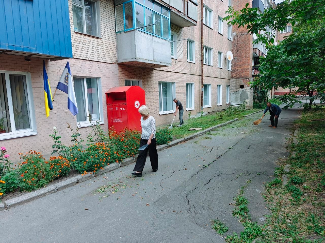 Три людини впорядковують та прибирають прибудинкову територію біля цегляного багатоповерхового будинку Координаційного центру «Вільні разом» у Хмельницькому під час екологічної акції до Всесвітнього дня прибирання «World Cleanup Day 2026», що організована для підтримання ладу та системної підтримки ВПО з Херсонщини. На передньому плані жінка у світлій блузі та чорних штанях іде асфальтованою доріжкою з совком у руці, а далі праворуч чоловік замітає асфальт великим віником, поки на задньому плані біля будівлі ще один працівник згрібає скошену траву та сміття. Ліворуч біля входу до простору на фасаді закріплено Державний Прапор України та прапор Херсонської області, поруч розбито квітник із помаранчевими чорнобривцями, а біля стіни встановлено великий червоний металевий бокс із логотипом Червоного Хреста для збору речей, що ілюструє спільну роботу колективу хабу із благоустрою прилеглої території.