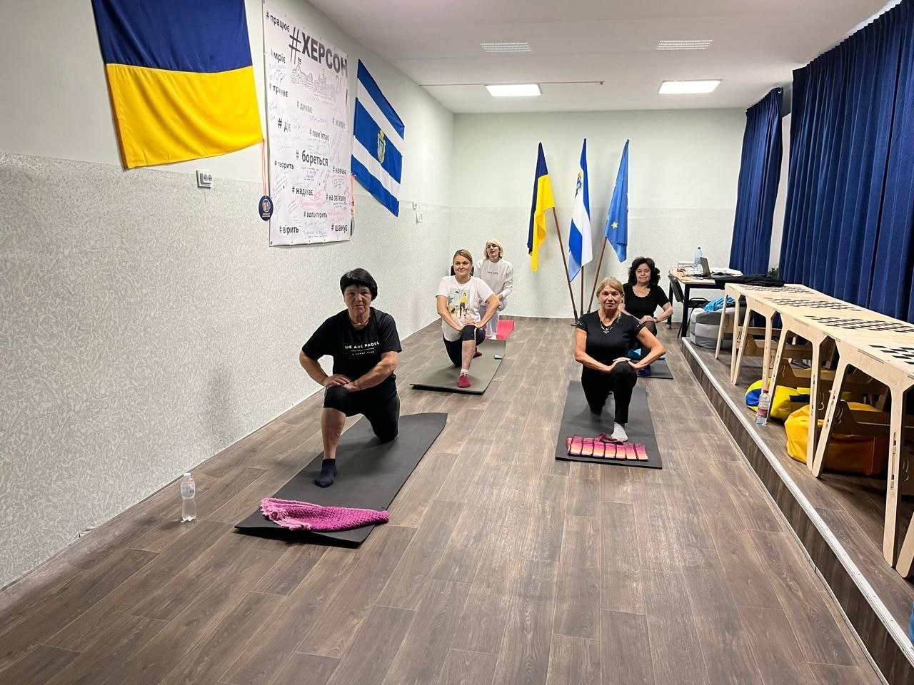 Група жінок різного віку займається на спортивних килимках у світлому залі Координаційного центру «Вільні разом» у Хмельницькому під час оздоровчого заняття з йоги, організованого в межах системної підтримки ВПО з Херсонщини. П'ятеро учасниць тренування вишикувалися у шаховому порядку на темних карематах, виконуючи вправу на розтяжку — глибокий випад уперед на одну ногу з опорою на коліно. Жінки вдягнені у зручний спортивний одяг і посміхаються в камеру. Заняття проходить у витягнутому приміщенні з ламінатом на підлозі, де праворуч вздовж темно-синіх штор облаштовано довгі дерев'яні столи, а на лівій стіні зали закріплено Державний Прапор України, прапор міста Херсона та великий плакат із написом «#ХЕРСОН» і патріотичними гаслами. На задньому плані на підставках додатково встановлено три прапори: України, Херсонської області та Європейського Союзу, що підкреслює спрямованість заходу на психоемоційне розвантаження та підтримку здоров'я переселенців.