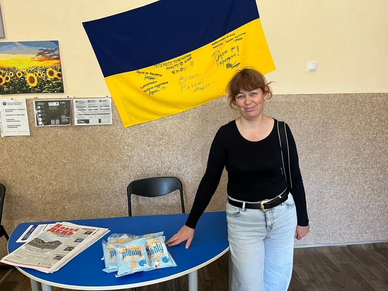 Жінка у чорній кофті з довгими рукавами та світло-блакитних джинсах стоїть у приміщенні біля синього напівкруглого столу, на якому розкладено пачки з рисовою сумішшю та стопку свіжих газет «Новий день» у межах системної підтримки ВПО з Херсонщини від Координаційного центру «Вільні разом» у Хмельницькому. Вона посміхається в камеру, тримаючи руку на краю столу з гуманітарною допомогою та друкованою пресою. На задньому плані на стіні висить великий синьо-жовтий Державний Прапор України з патріотичними написами від руки та автографами «Вільні Разом!», а ліворуч — інформаційні листки та картина із соняшниковим полем.