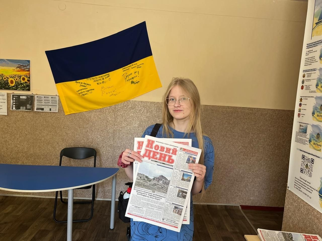 Дівчина в окулярах та синій сукні стоїть у приміщенні Координаційного центру «Вільні разом» у Хмельницькому та тримає перед собою розгорнутий примірник обласної газети «Новий день», що відображає системну інформаційну та гуманітарну підтримку ВПО з Херсонщини. Вона посміхається в камеру, демонструючи першу шпальту друкованого видання з великим червоним заголовком. Ліворуч від неї розташований порожній синій напівкруглий стіл із чорним стільцем, а на задньому плані на світло-жовтій стіні висить Державний Прапор України з патріотичними написами від руки та автографами. Справа на стіні закріплено великий вертикальний інформаційний плакат із логотипами хабу, картою та QR-кодами, що ілюструє всебічну допомогу людям, які змушені були залишити свої домівки через війну.