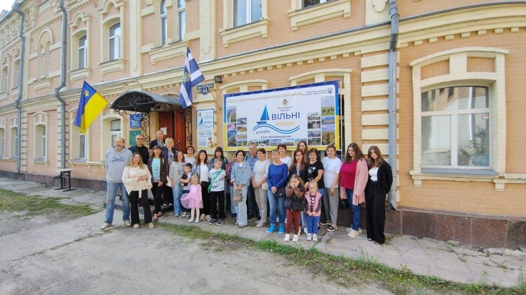 Велика група переселенців з Херсонщини разом із працівниками осередку урочисто позує для спільного фото на вулиці біля будівлі Координаційного центру «Вільні разом» у Кропивницькому, де в межах всебічної підтримки провели науково-пізнавальний захід «Зоопарк під мікроскопом», майстер-клас «Солодкий Херсон», конкурс малюнків та кіноперегляд. Понад шістдесят дітей і дорослих, які долучилися до масштабної програми заходів, вишикувалися в кілька рядів на тротуарі на тлі двоповерхового цегляного будинку бежевого кольору. Позаду них на фасаді закріплено великий офіційний білий банер із синім логотипом хабу, написом «ВІЛЬНІ РАЗОМ для ВПО місто Кропивницький» та фотоілюстраціями херсонських краєвидів, а ліворуч над входом під кованим дашком майорять Державний Прапор України та синьо-білий прапор міста Херсона.
