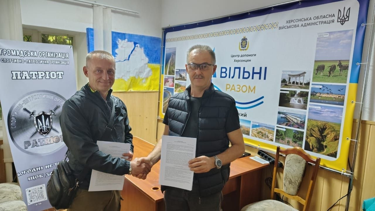 Двоє чоловіків стоять у кабінеті Координаційного центру «Вільні разом» у Кропивницькому та потискають один одному руки на знак укладання меморандумів про співпрацю з ГО «Спортивно-патріотична організація «Патріот» та БО «100% життя. Кропивницький» для всебічної підтримки переселенців з Херсонщини. Ліворуч зафіксовано представника організації у чорній куртці, а праворуч — чоловіка в окулярах та чорній дутій жилетці, який тримає підписаний офіційний документ. Вони позують на тлі великого брендованого банера під егідою Херсонської обласної військової адміністрації з написом «Центр допомоги Херсонцям ВІЛЬНІ РАЗОМ» та колажем із херсонськими краєвидами, а ліворуч біля вікна встановлено інформаційний стенд організації «ПАТРІОТ» із символікою тризуба.