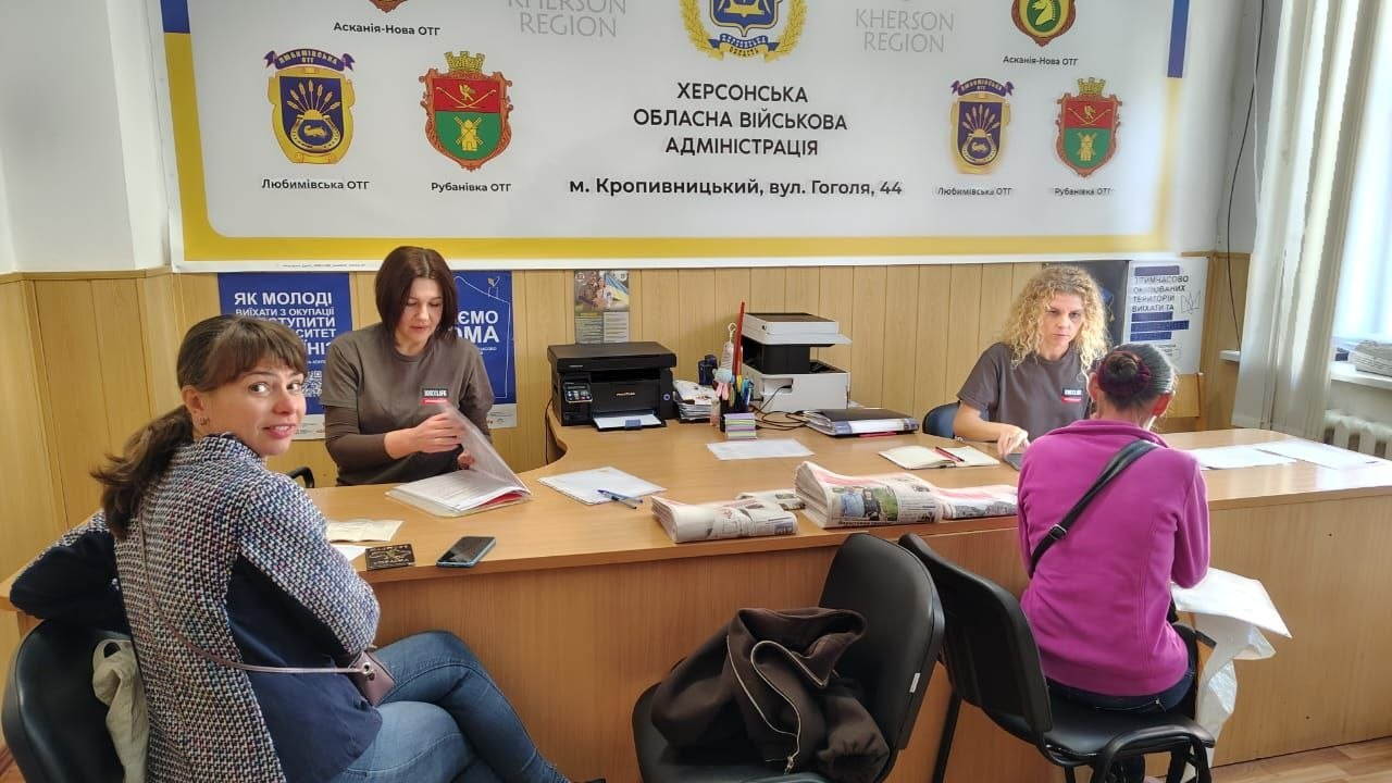 Дві працівниці Координаційного центру «Вільні разом» у Кропивницькому за великим кутовим столом приймають та консультують відвідувачів у межах всебічної підтримки переселенців з Херсонщини та реалізації проєкту «Картка Херсонщини». На передньому плані ліворуч на стільці сидить жінка у сірому жакеті, яка повернула голову до камери, а праворуч спиною до глядача зафіксовано відвідувачку у рожевій кофті, яка спілкується зі співробітницею хабу. За столом працюють дві працівниці у сірих фірмових футболках, перед якими розкладено документи, бланки реєстрації та примірники обласної газети «Новий день», що видаються землякам. На задньому плані встановлено великий білий банер із гербами громад та підписом «Херсонська обласна військова адміністрація».